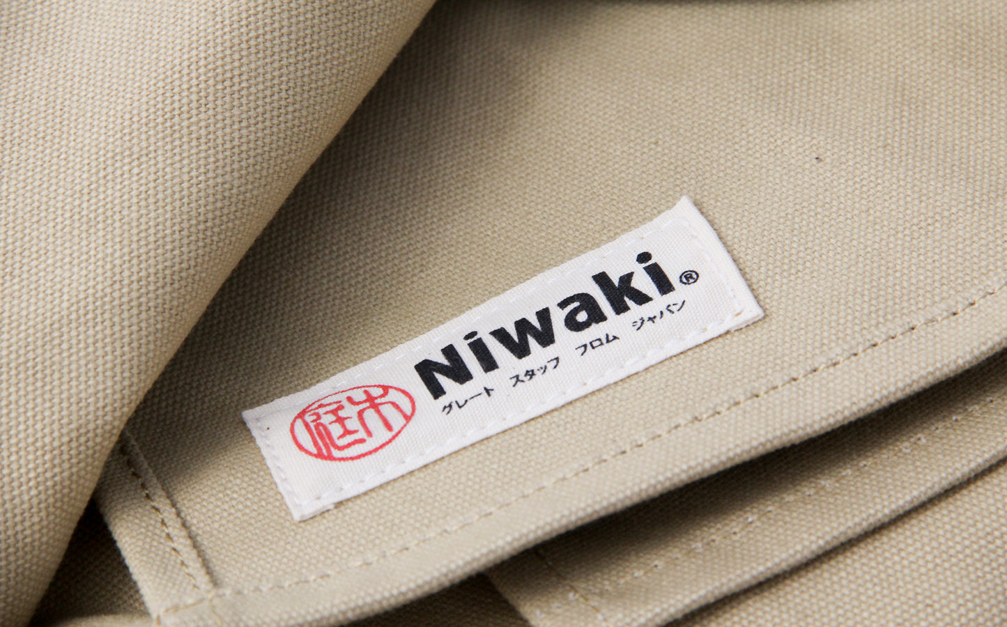 Eine Nahaufnahme des beigen Stoffes der Niwaki | Schürze „Split Apron“ (94 x 63 cm) zeigt ein weißes Etikett mit der Aufschrift „Niwaki“ in schwarzer Schrift und japanischen Schriftzeichen, ergänzt durch ein rotes, rundes Logo – ein Zeichen für die hochwertige Verarbeitung, die man von dieser Premium-Gartenschürze „Canvas“ von Niwaki erwartet.