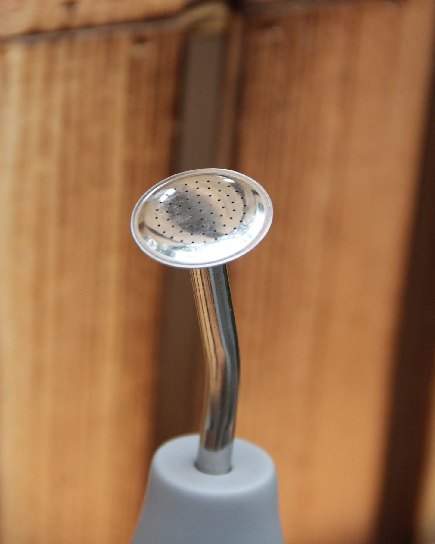 Eine Nahaufnahme der „Esschert Gießbrause Ball Grau L“, einer Gießkanne aus Metall mit kleinen Löchern für eine wassersparende und gezielte Pflanzenbewässerung, die an einem weißen Griff befestigt ist und vor einem Holzhintergrund steht.