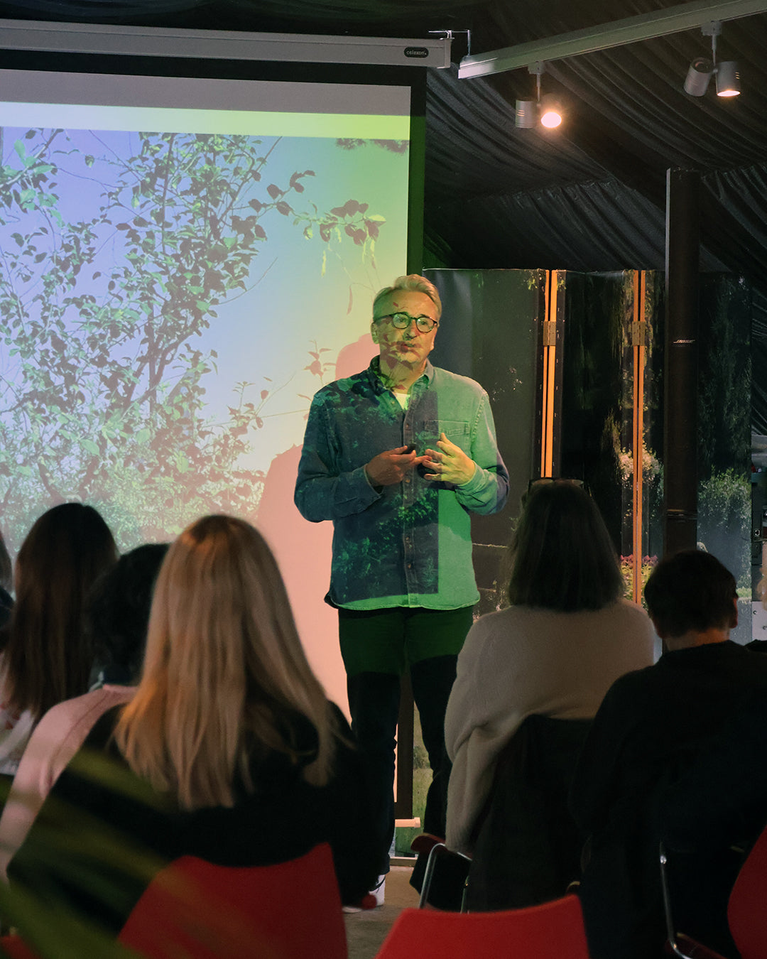 Horst Mager gibt beim HALBTAGESKURS "Das Gartenjahr-Monat für Monat" der Königlichen Gartenakademie am Fr. 13.03.2026, 13-16 Uhr, vor einem aufmerksamen Publikum in roten Stühlen fachkundige Gartentipps, während hinter ihm Naturbilder projiziert werden.