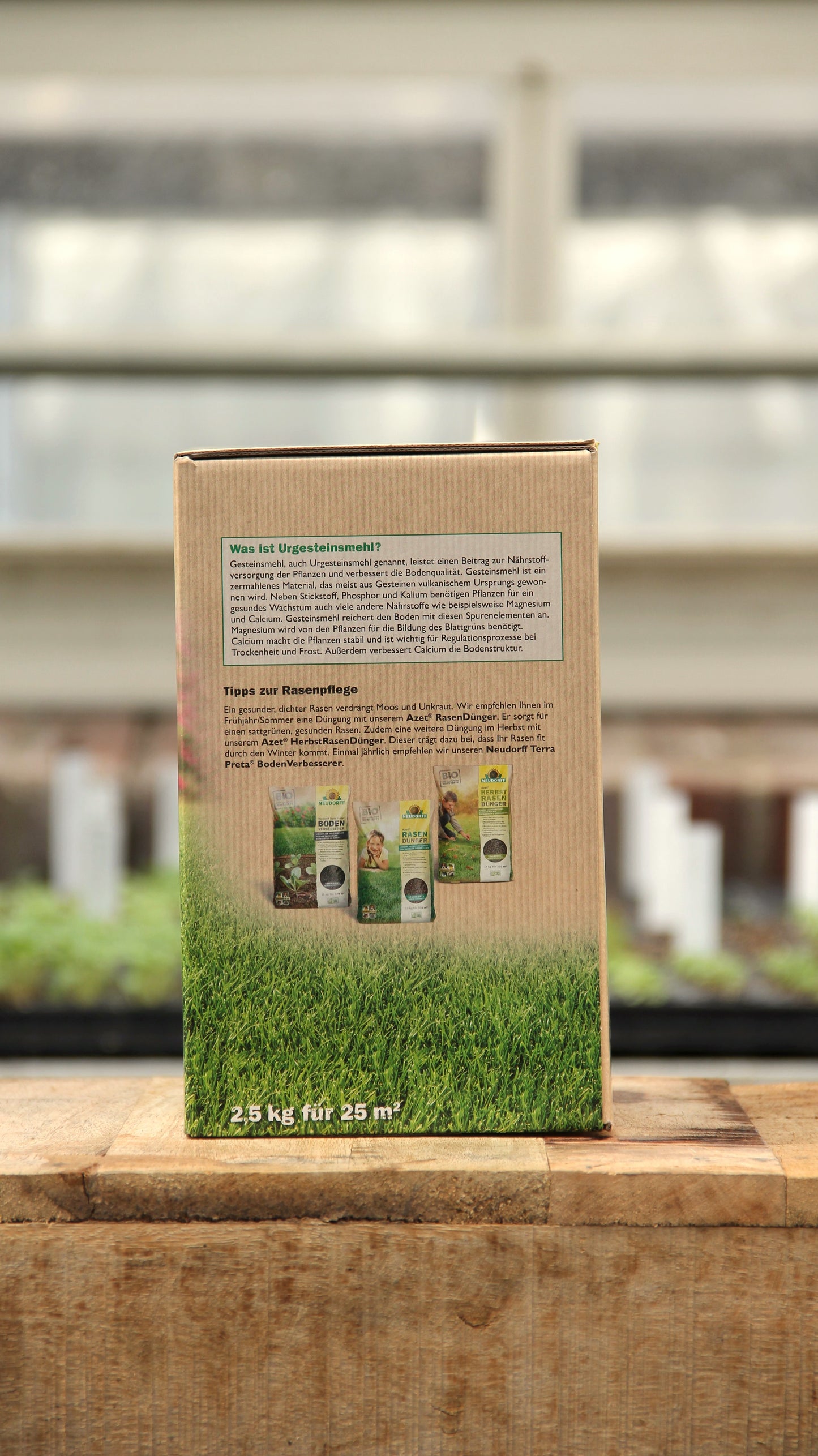 Ein Neudorff Urgesteinsmehl aus Diabasgesteinsmehl vulkanischen Ursprungs 2,5 kg Pappkarton mit deutschem Text steht auf einer Holzfläche an einem Fenster, Grünpflanzen verschwimmen im Hintergrund.
