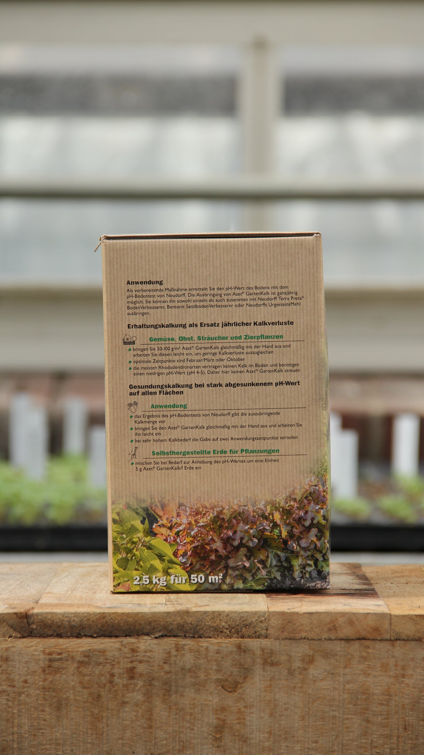 Eine Neudorff-Box mit der Aufschrift "Gartenkalk Bodenverbesserer aus natürlichen Rohstoffen 2,5 kg" mit Abbildungen von Blattpflanzen steht auf Holz, ideal zur Regulierung des pH-Werts des Bodens; im Hintergrund sind verschwommene Gewächshausregale zu sehen.