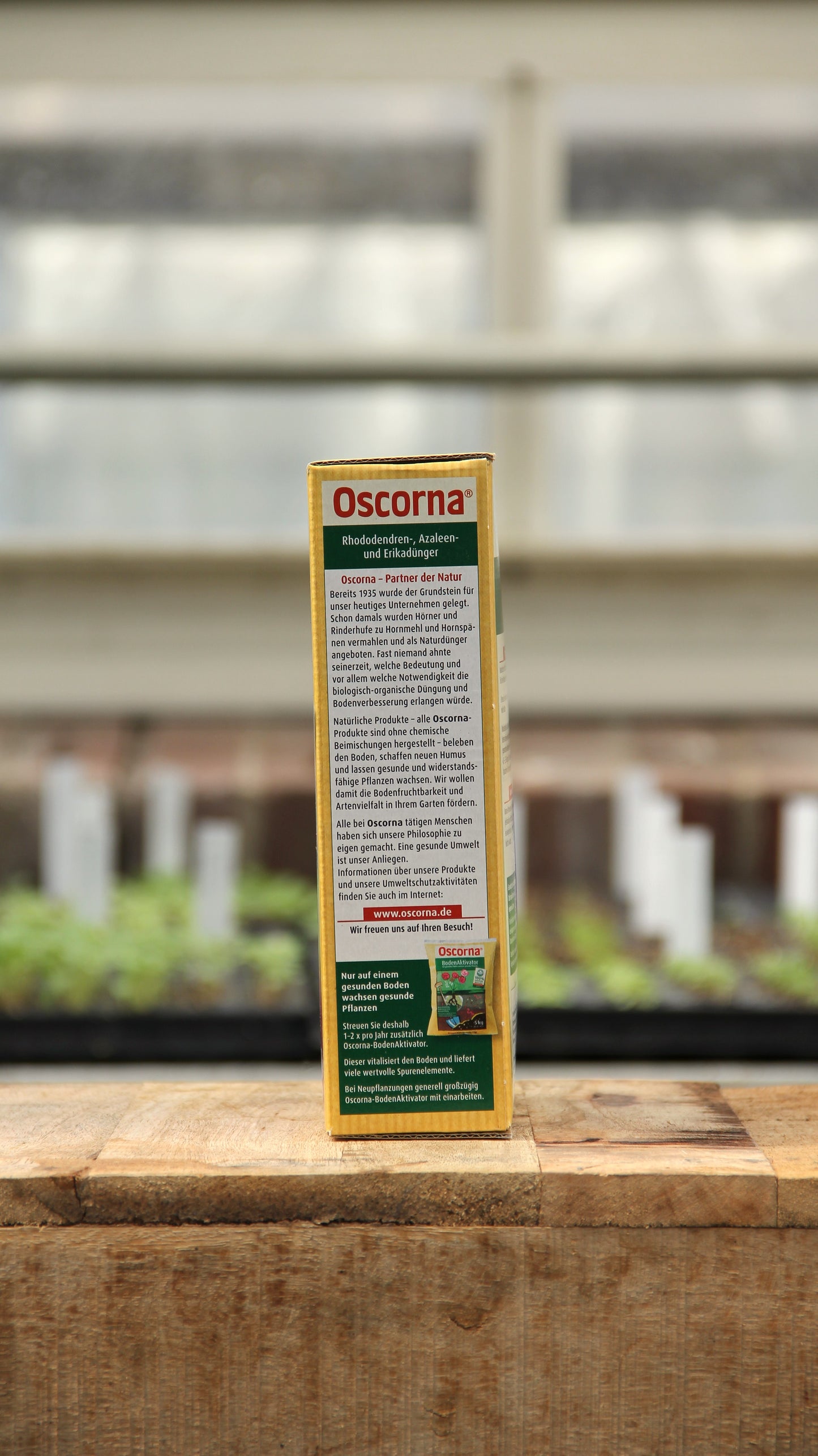 Eine Schachtel Oscorna | Rhododendren-, Azaleen- und Erikadünger Naturdünger 1 kg steht auf einer Holzfläche mit unscharfem pflanzlichem Hintergrund; das deutsche Etikett ist sichtbar.