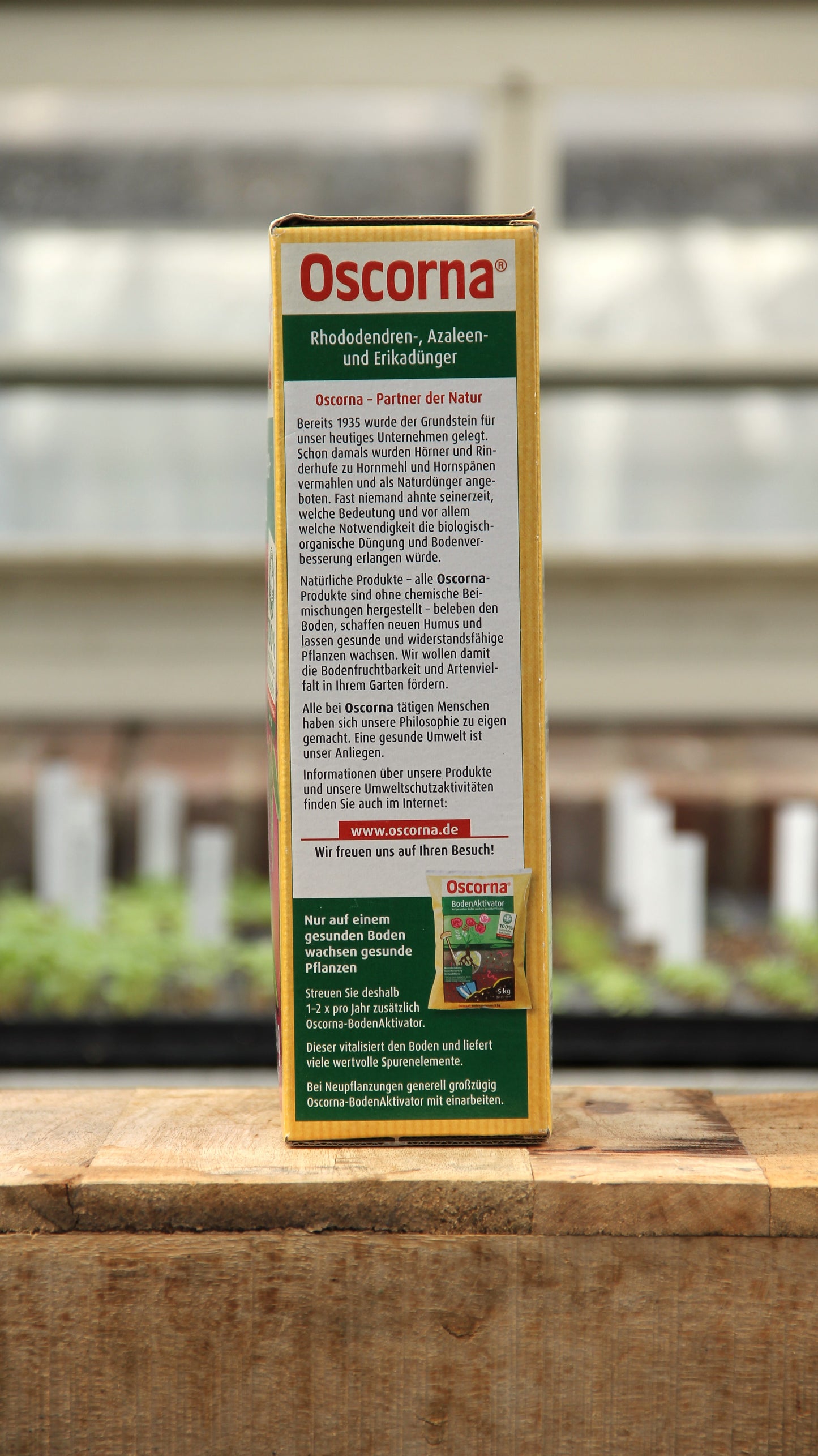 Eine Kiste Oscorna Rhododendren-, Azaleen- und Erikadünger Naturdünger 2,5 kg steht auf einer Holzfläche, auf der Produkttexte und -abbildungen zu sehen sind; im Hintergrund sind unscharfe Gewächshausregale mit Moorbeetpflanzen zu sehen.
