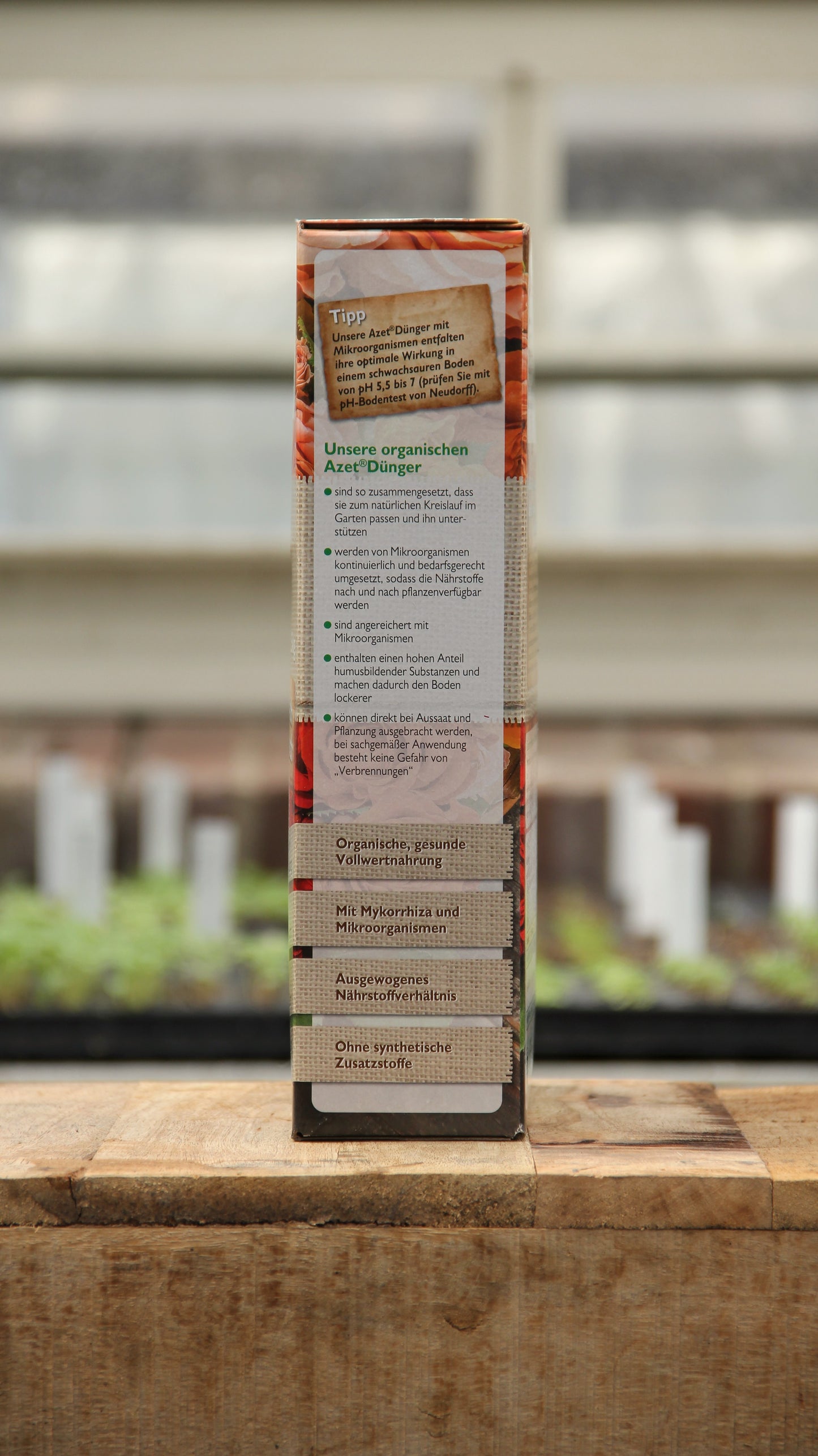 Seitenansicht einer Neudorff | Azet Rosen-Dünger 2,5 kg Kiste auf einer Holzoberfläche, mit deutschem Produktvorteilstext. Der Hintergrund zeigt unscharfe Pflanzen und Pflanzschalen in einem Gewächshaus.