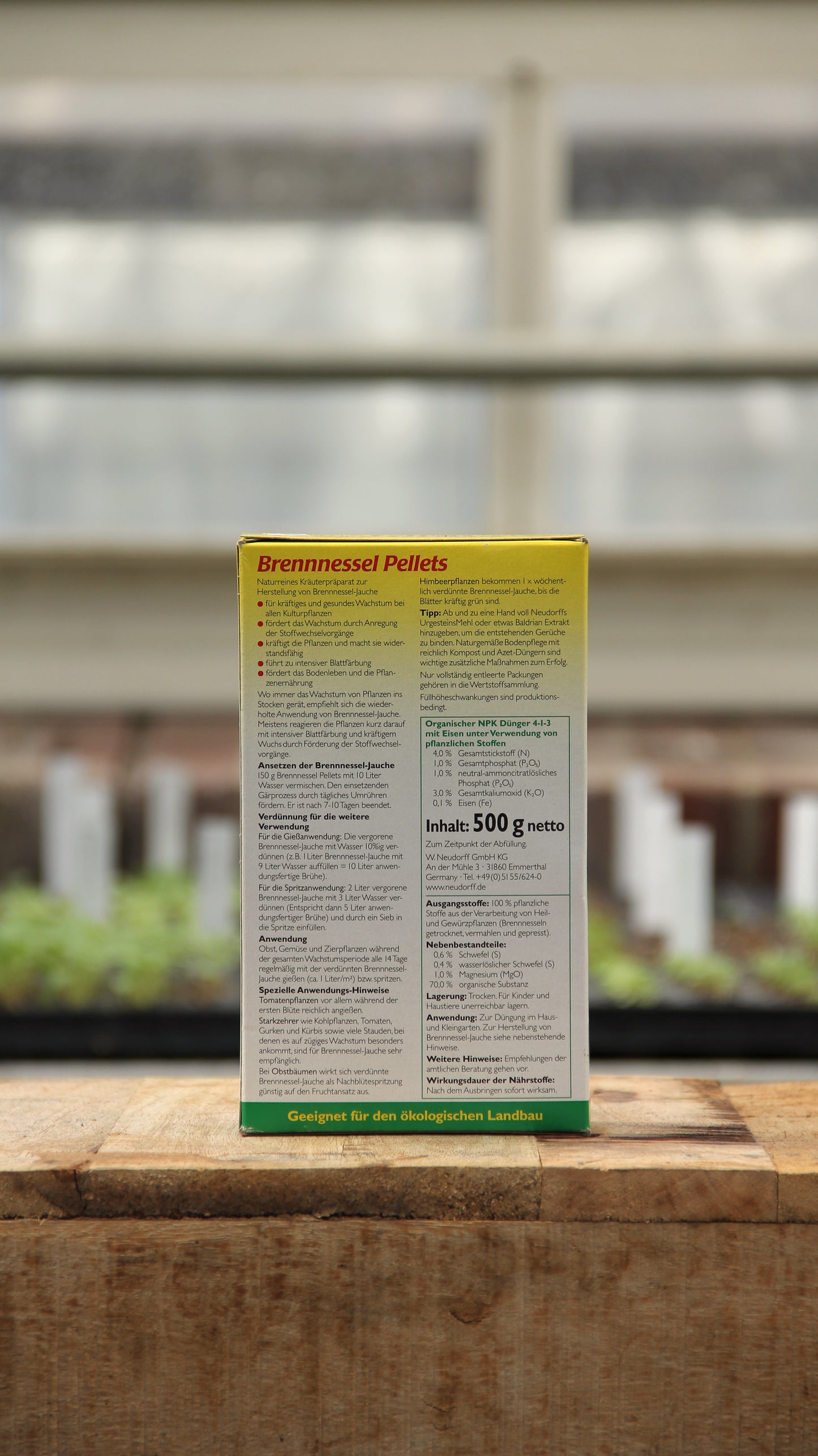 Eine 500-Gramm-Packung Neudorff | Brennnessel Pellets organischer Dünger steht auf Holz und zeigt auf der Rückseite Tipps für den deutschen Ökolandbau. Im unscharfen Hintergrund sind junge Setzlinge und ein Gewächshausfenster zu sehen.