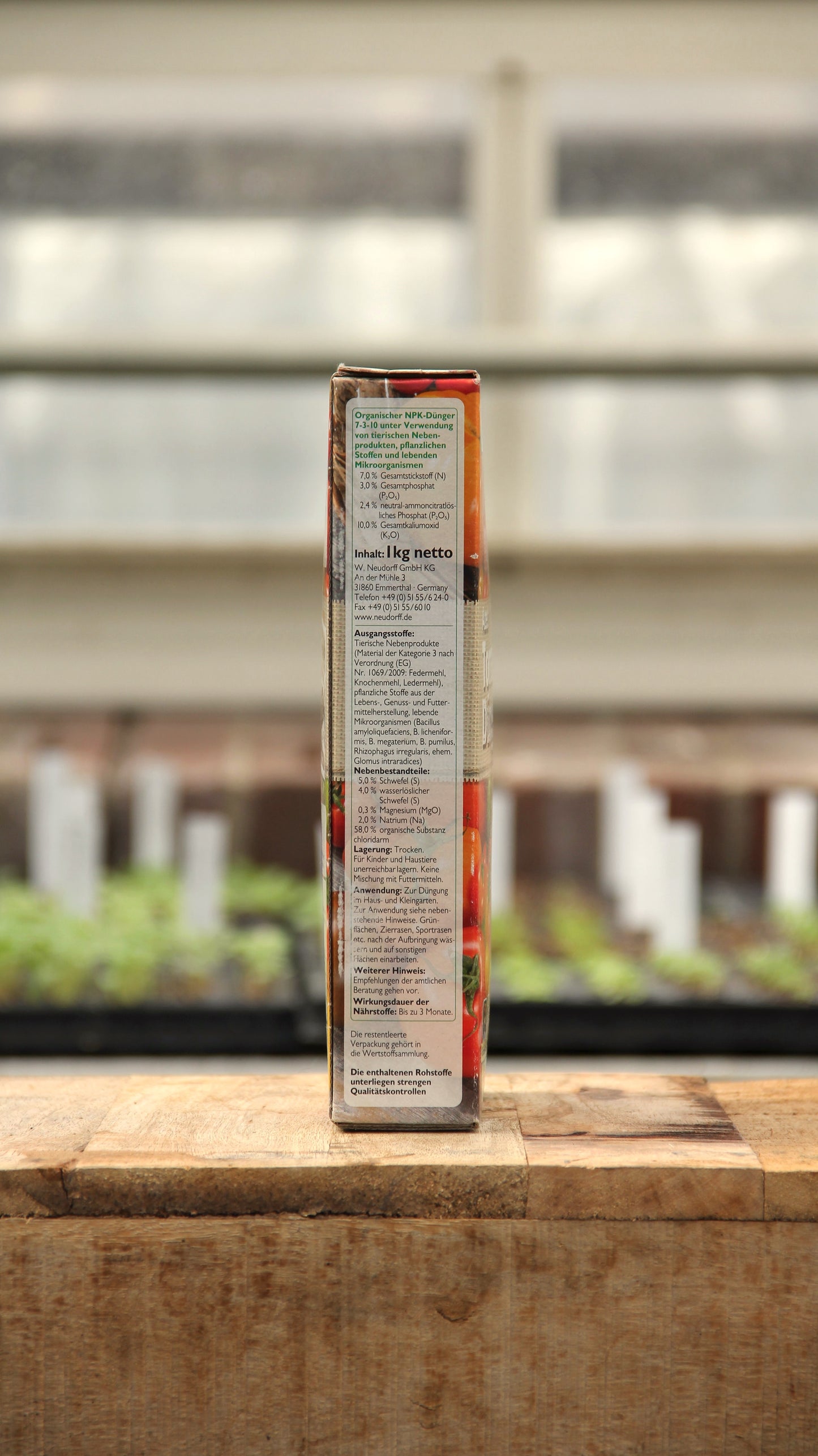 Eine Seitenansicht der Neudorff Azet Tomaten-Dünger 1 kg-Verpackung zeigt die Nährwertangaben und das Etikett mit den Inhaltsstoffen auf einer Holzfläche mit unscharfem Gewächshaushintergrund.