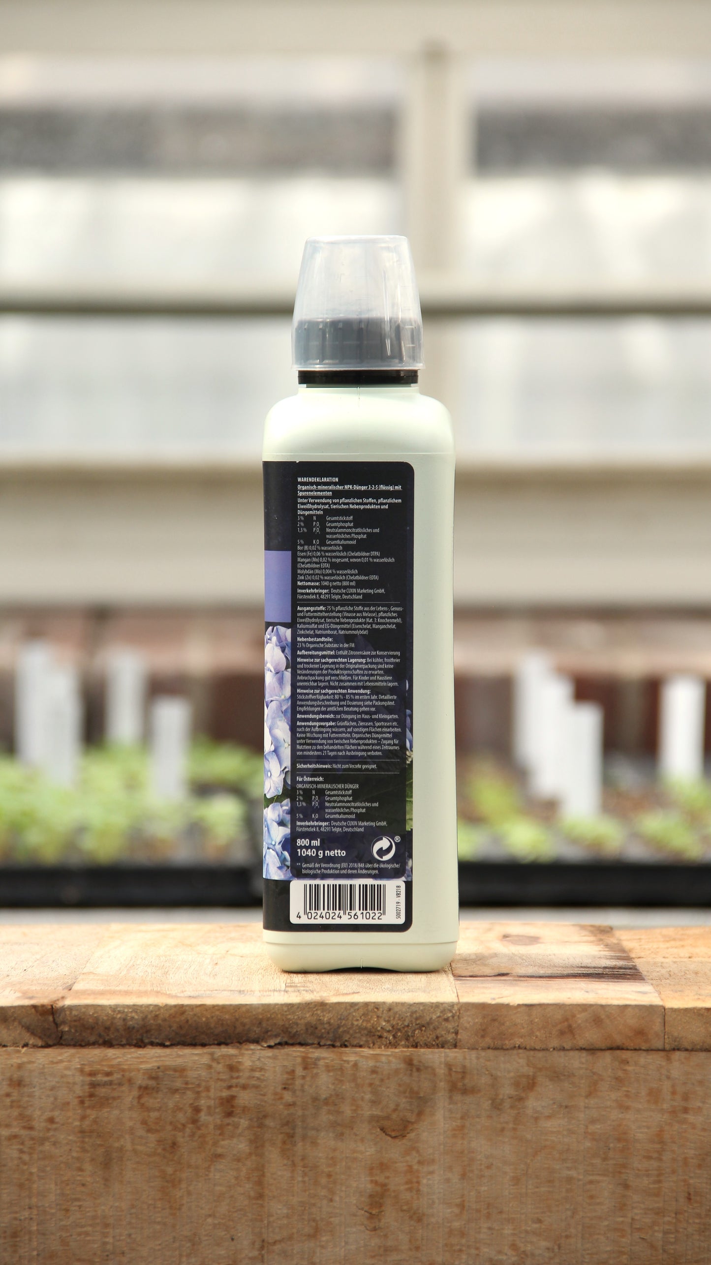 Eine weiße 800-ml-Plastikflasche Cuxin Rhododendren- und Hortensien Flüssigdünger mit klarem Verschluss steht auf Holz, das Rückenetikett zeigt Text und Blumen. Unscharfer Gewächshaushintergrund - ideal für Moorbeetpflanzen.