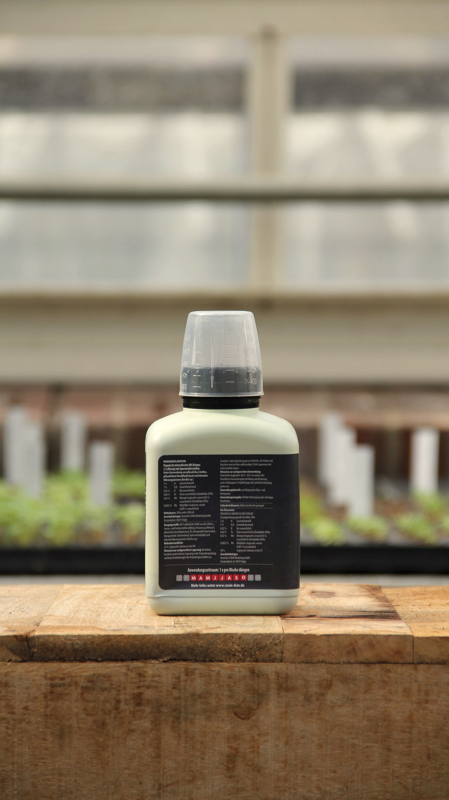 Eine 400-ml-Flasche Cuxin | Flüssigdünger Mediterrane Pflanzen & Zitruspflanzen Bio mit schwarzem Etikett und klarem Verschluss steht auf Holz vor einem unscharfen Gewächshaus, ideal für Zitrus- und mediterrane Pflanzen. Der weiße Etikettentext ist unleserlich.