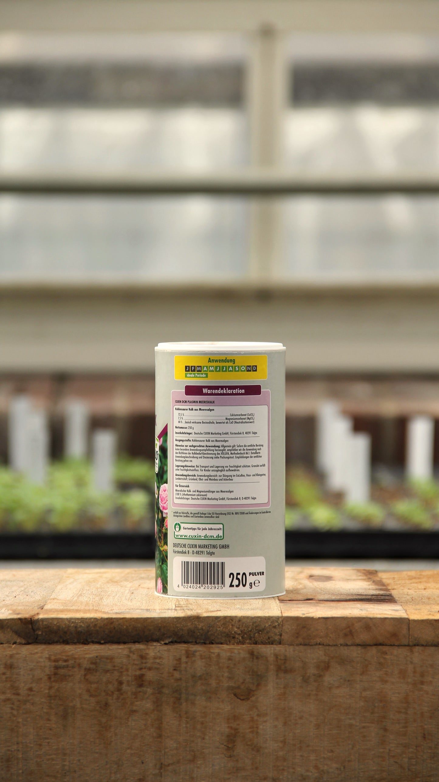 Ein Behälter mit Cuxin | Plasmin Meereskalk 250 g von der Königlichen Gartenakademie steht auf einer Holzfläche in einem Gewächshaus, das Etikett und die Gebrauchsanweisung sind sichtbar; im Hintergrund sind unscharfe Setzlinge und Pflanzenmarker zu sehen.