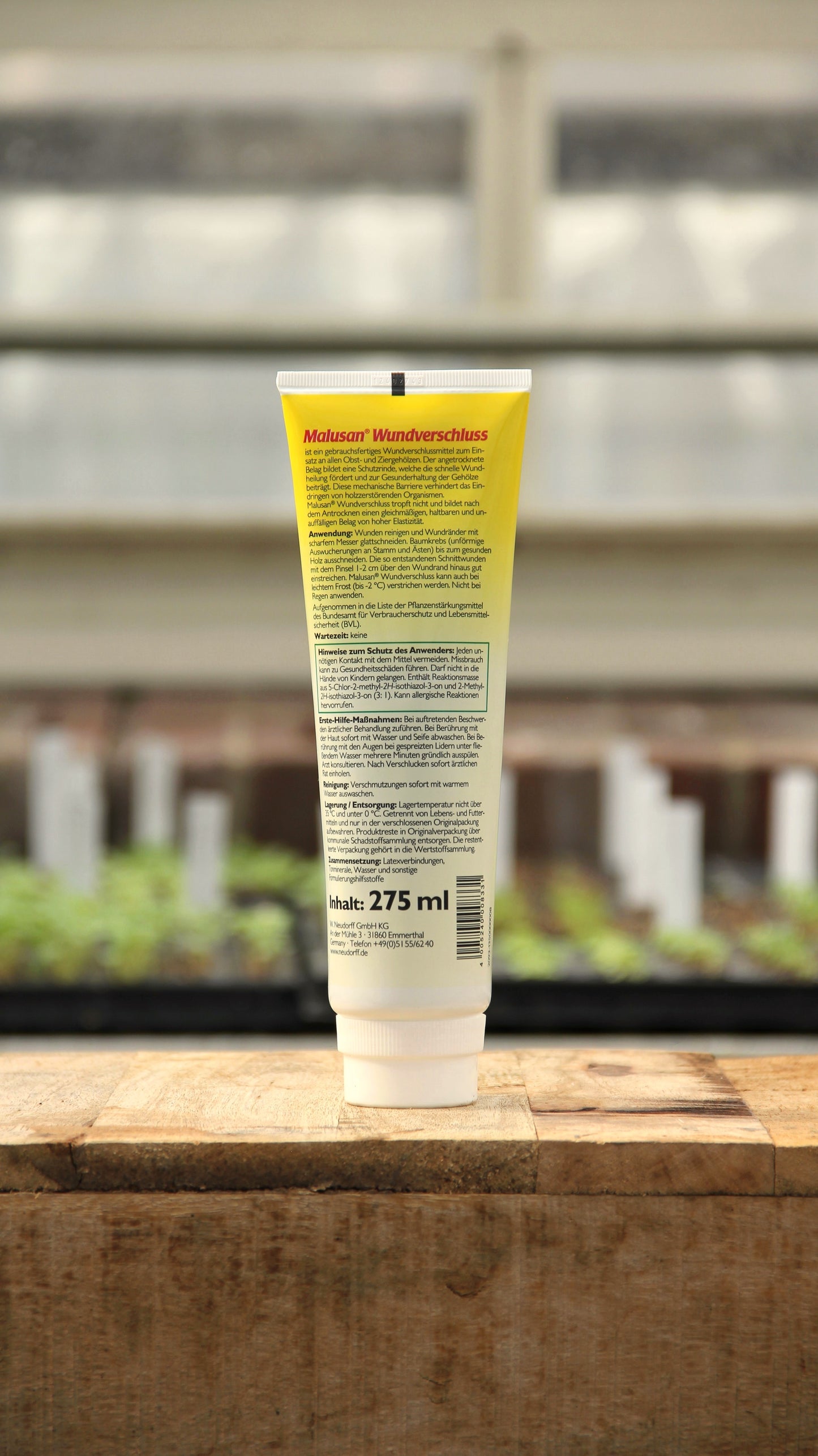 Eine 275-ml-Tube Neudorff | Malusan Baumwundverschluss mit gelbem und weißem Etikett steht aufrecht auf einer Holzfläche mit unscharfen Pflanzen und einem Gewächshausfenster im Hintergrund.