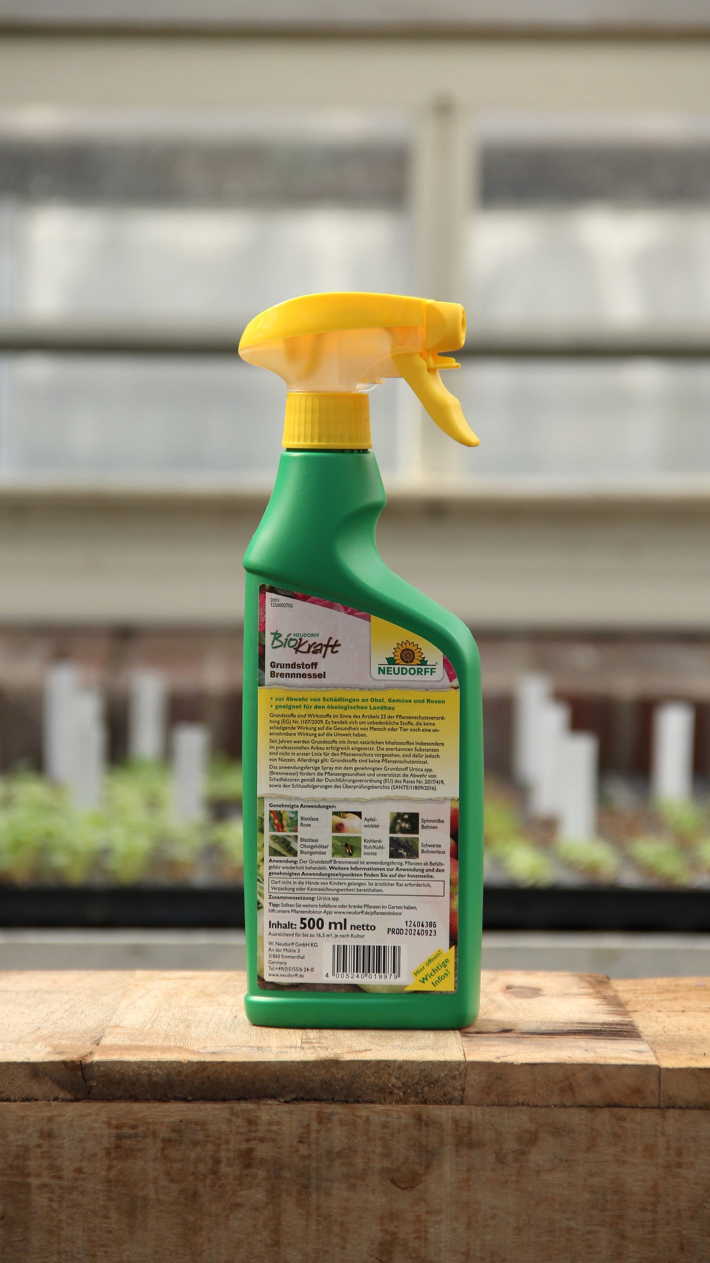 Eine grüne Neudorff BioKraft Grundstoff Brennnessel 500 ml Sprühflasche mit gelbem Abzug steht auf Holz, Rückenetikett sichtbar, inmitten unscharfer Gewächshauspflanzen - ideal für natürlichen Pflanzenschutz gegen Blattläuse.
