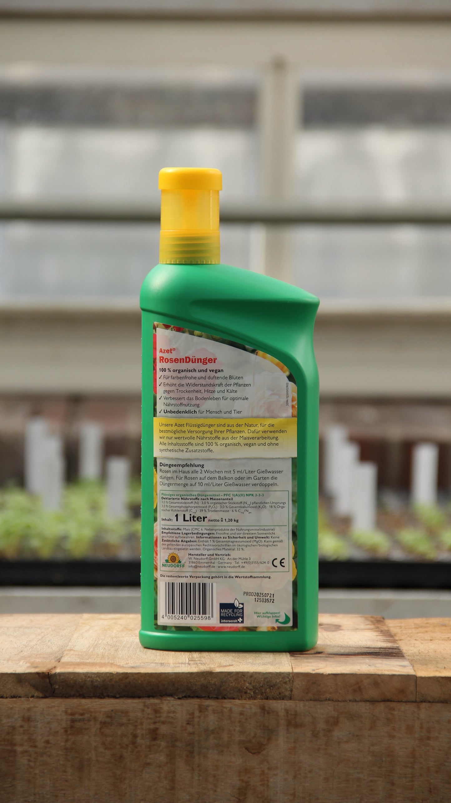 Eine grüne Kunststoffflasche Neudorff Azet Rosendünger flüssig 1 l mit gelbem Verschluss steht auf Holz. Im Hintergrund sind Setzlinge und ein unscharfes Gewächshausfenster zu sehen; das Etikett ist überwiegend mit roter und schwarzer deutscher Schrift versehen.