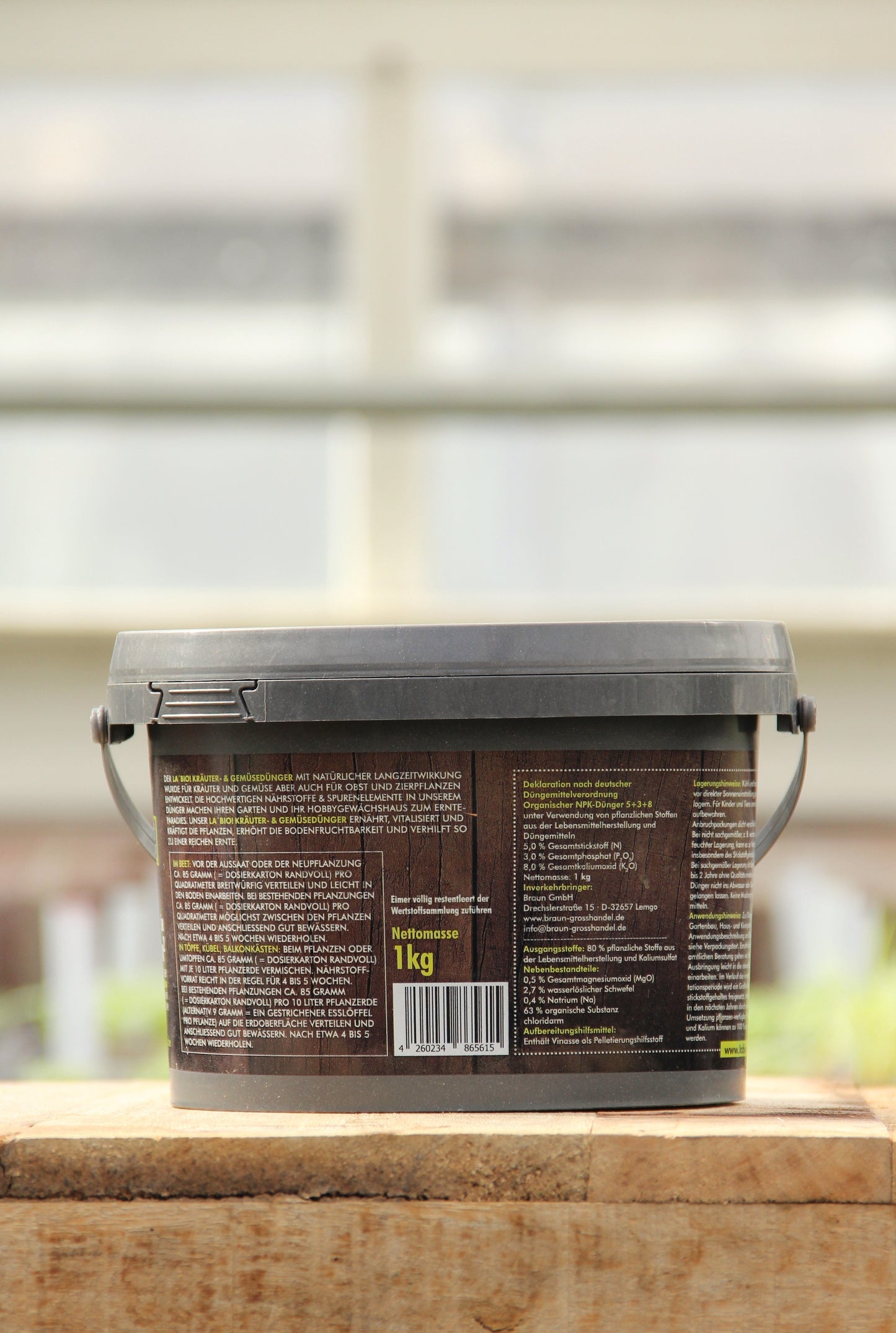 Ein 1 kg La'Bio | Kräuter- & Gemüsedünger Kunststoffbehälter mit schwarzem Deckel und Henkel, braunem mehrsprachigem Etikett, veganer Kennzeichnung, ruht auf Holz mit einem verschwommenen Innenhintergrund.