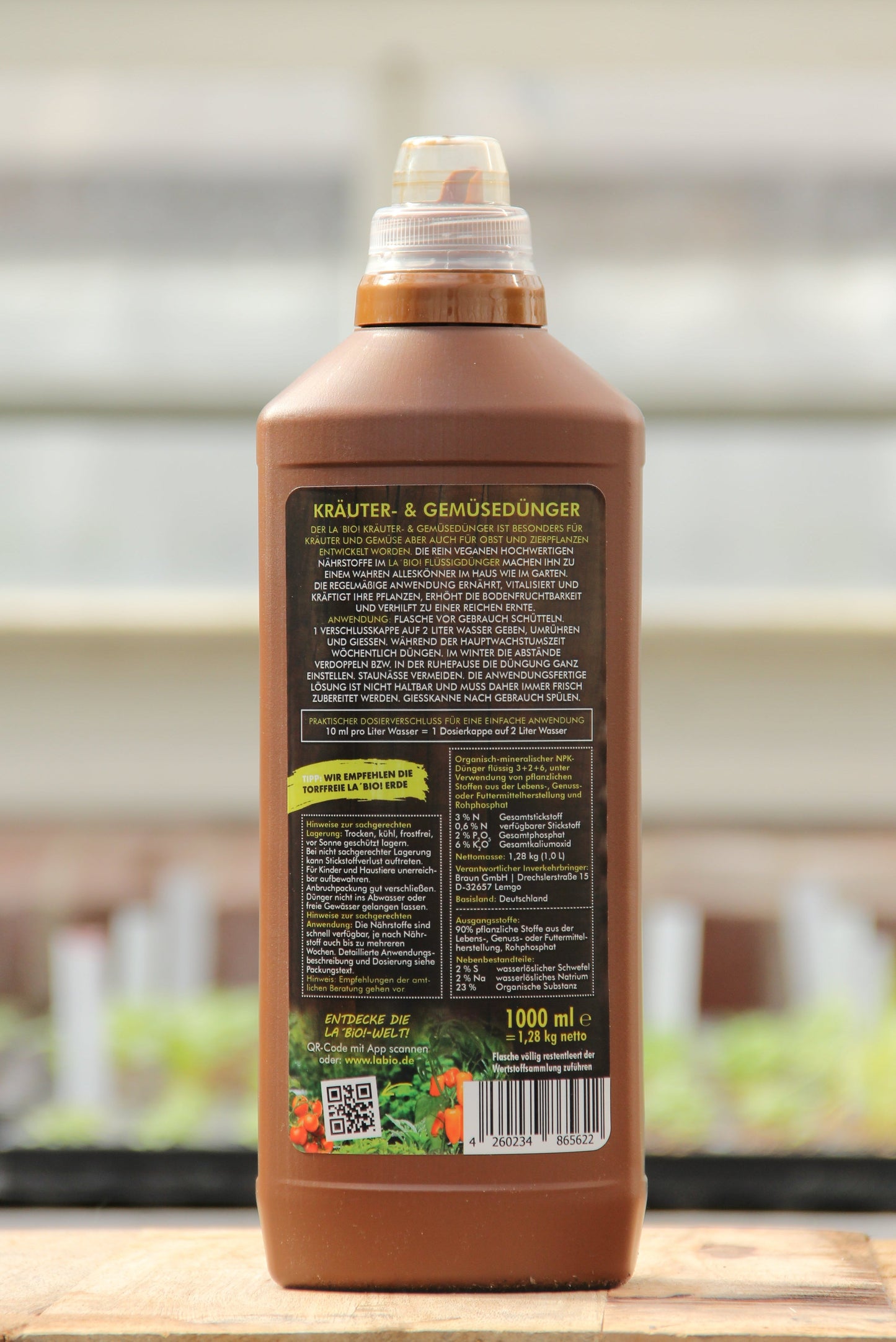 Eine braune Plastikflasche La'Bio | Kräuter & Gemüsedünger 1 l von LaBio mit braunem Verschluss, von hinten gesehen. Das deutsche Etikett zeigt Anweisungen und Dosierungstabellen. Im unscharfen Hintergrund sind grüne Pflanzen zu sehen.