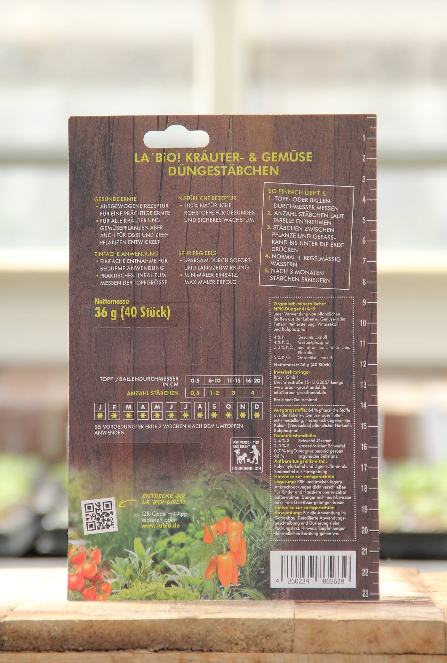 Rückseite der La'Bio Düngestäbchen 40 Stk. Packung von La'Bio, mit deutscher Anleitung, Dosierungstabelle, Strichcode und Abbildungen von Tomaten und Kräutern für gesundes Wachstum. Der Hintergrund zeigt unscharfe Zimmerpflanzen und ein Fenster.