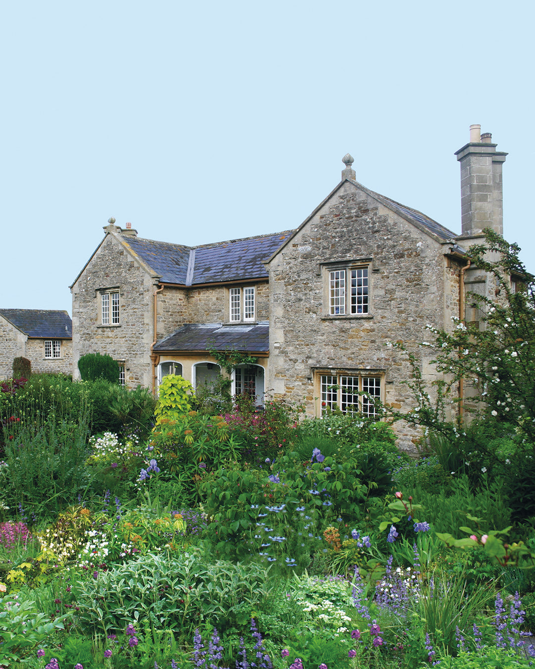 Ein steinernes Häuschen mit Schieferdach steht in einem lebendigen Garten und erinnert an "Der Cottage Garten: Der Garten der Zukunft", ein Halbtageskurs der Königlichen Gartenakademie am Sa. 07.03.2026, wo unter strahlend blauem Himmel das Gärtnern der Zukunft gelehrt wird.