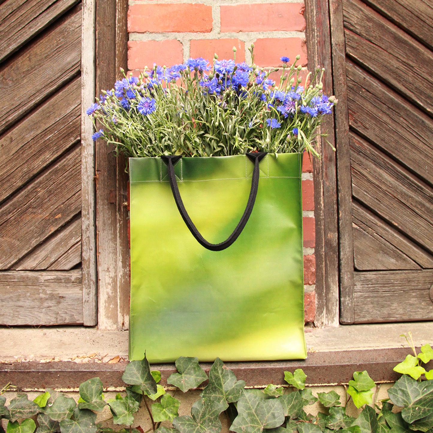 Die Upcycling Tasche 'Leaf' aus Werbeplane von Königliche Gartenakademie steht auf einer Fensterbank mit lila Blumen vor einer Backsteinmauer mit hölzernen Fensterläden, während darunter grüner Efeu wächst.