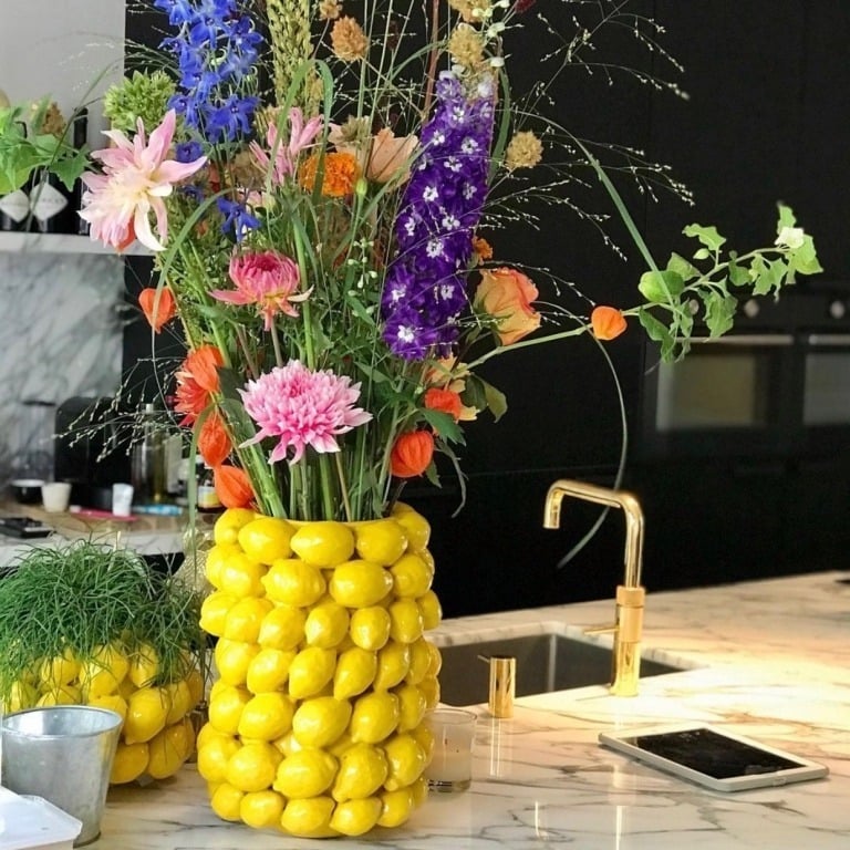 Ein bunter Blumenstrauß steht in einem Des Pots | Blumentopf schmal mit Zitronen in gelb H 16 Ø 18 cm von Des Pots auf einer Küchenarbeitsplatte aus Marmor, neben einem goldenen Wasserhahn, einer kleinen Pflanze und einem Tablet.