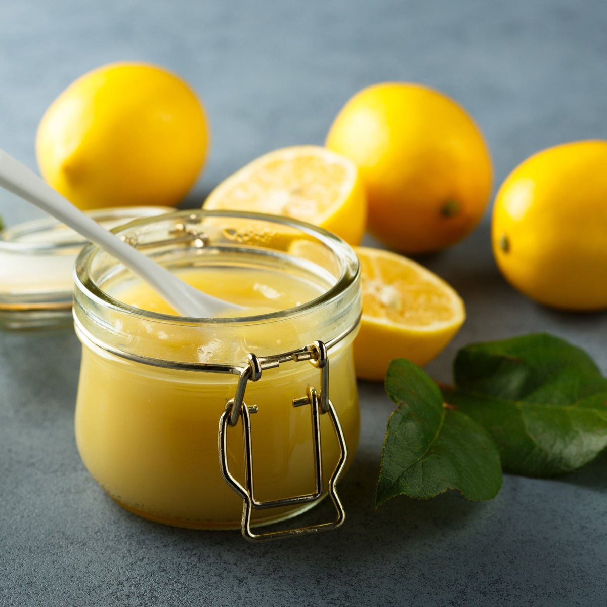 Ein 312 g schweres Glas von Wilkin & Sons | Tiptree Lemon Curd mit einem weißen Löffel darin steht auf einer grauen Unterlage, umgeben von ganzen Zitronen, Zitronenhälften und grünen Blättern.