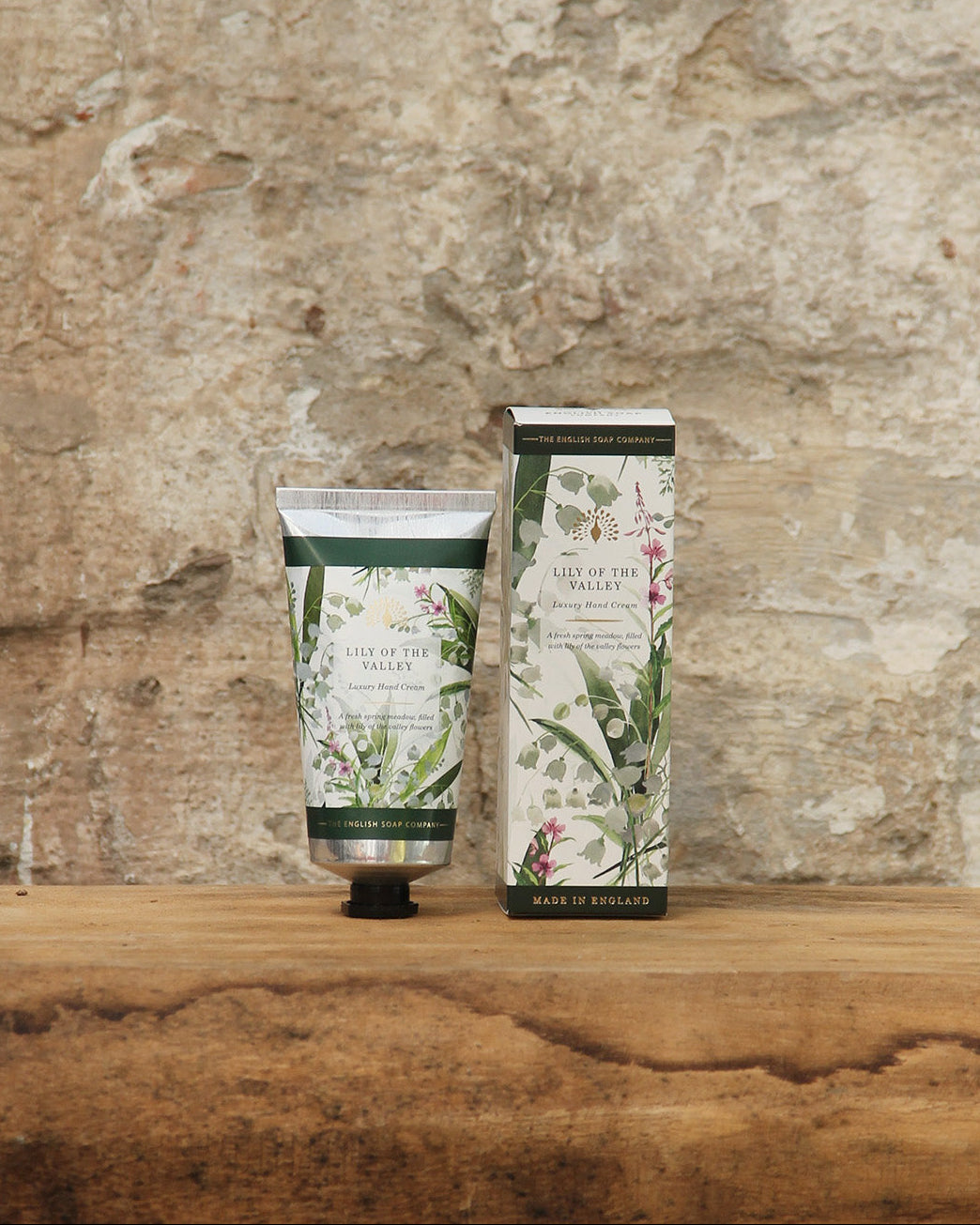 Eine Tube The English Soap Company ESC | 'Lily of the Valley' Handcreme mit Maiglöckchenduft 75 ml ruht neben der dazugehörigen Schachtel auf einer rustikalen Holzfläche mit einer strukturierten Steinwand im Hintergrund.