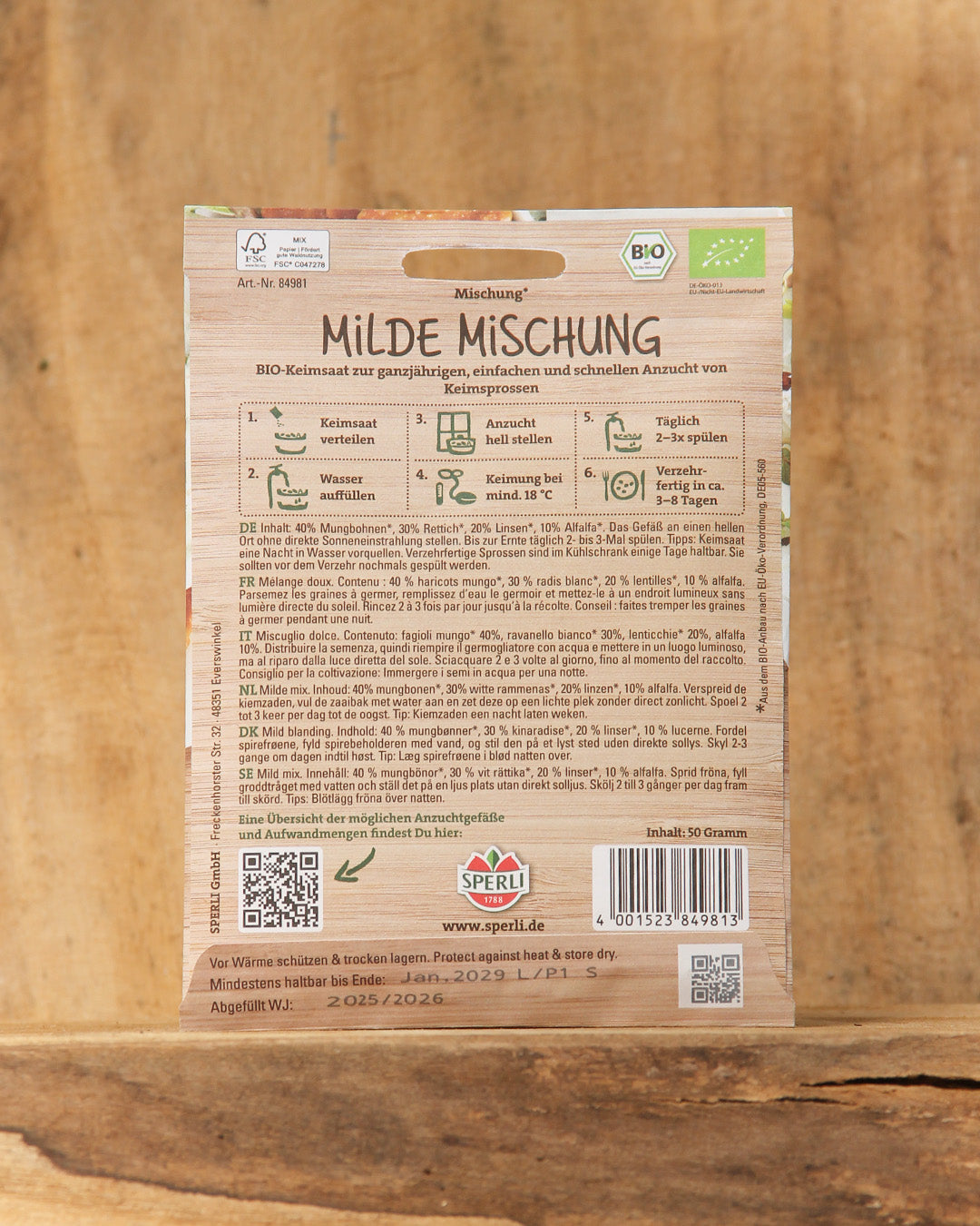 Eine Packung Sperli's | Bio Keimsprossen Saatgut 'Milde Mischung' wird auf einer Holzfläche präsentiert. Die Verpackung enthält eine Anbauanleitung, einen QR-Code, Bio-Zertifizierungssymbole und detaillierte Informationen - ideal für den Sprossenanbau zu Hause.