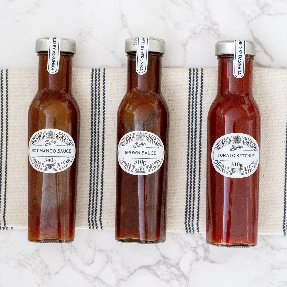 Drei Glasflaschen mit Soßen – „Wilkin & Sons Tiptree Steak Sauce Rauchig würzig“, „typisch englische Würzsauce (Brown Sauce)“ und Tomatenketchup – stehen in einer Reihe auf einem gestreiften Tuch, das auf einer weißen Marmorplatte liegt.