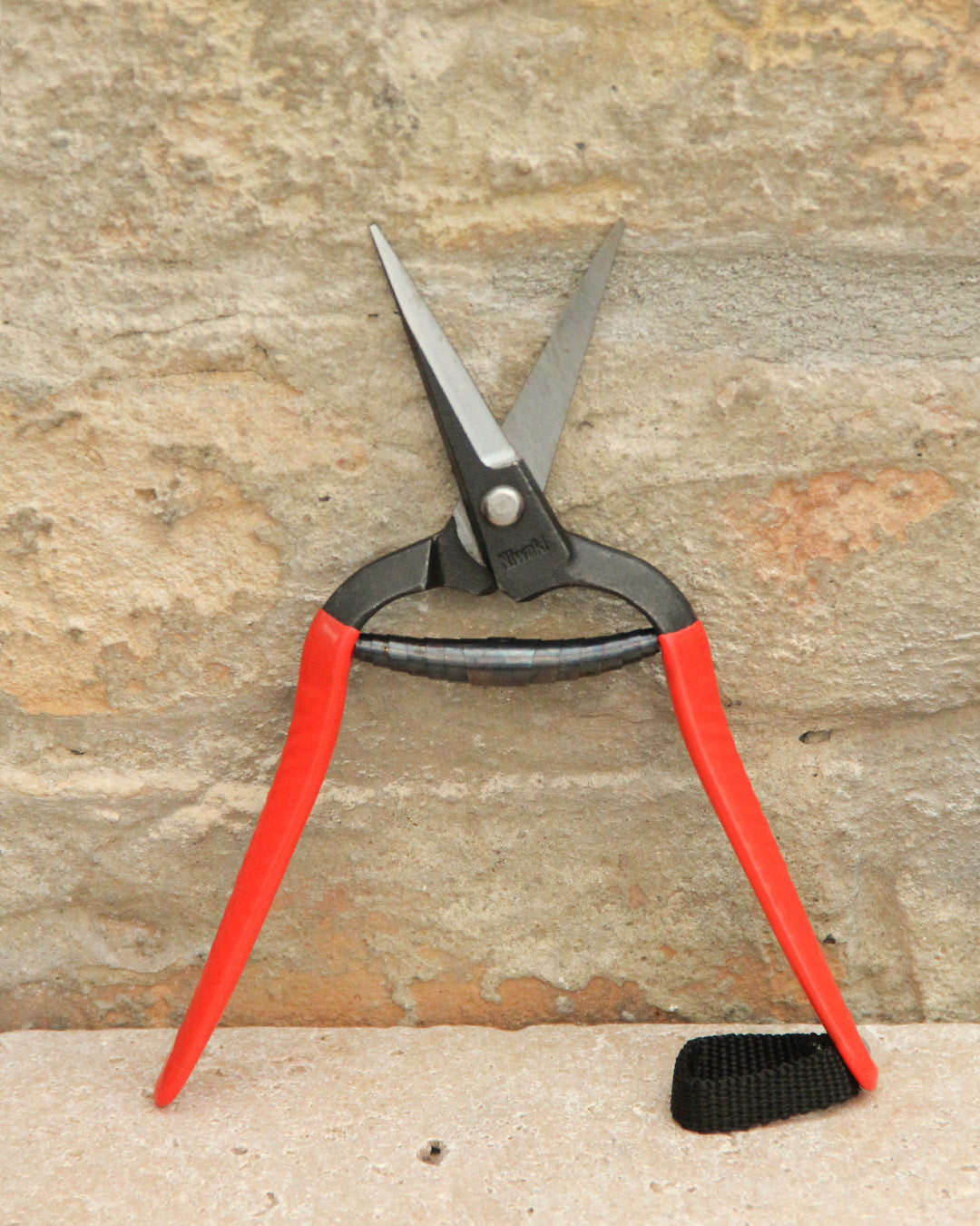 Ein Paar Niwaki | 'Forged Snips' Blumenschere von Niwaki mit roten Griffen und einer offenen Klinge steht aufrecht vor einer strukturierten Steinwand.