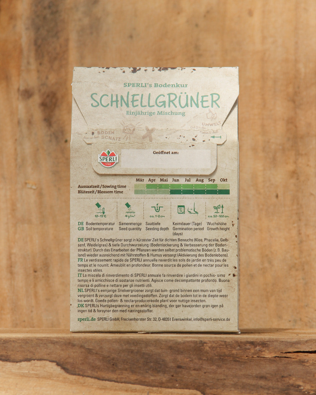 Eine 450g-Papiertüte der Sperli's Bodenkur 'Schnellgrüner' steht auf einer Holzunterlage und zeigt auf der Rückseite Aussaat- und Blütezeiten, Pflanzhinweise und deutsche Symbole für die optimale Bodenverbesserung.