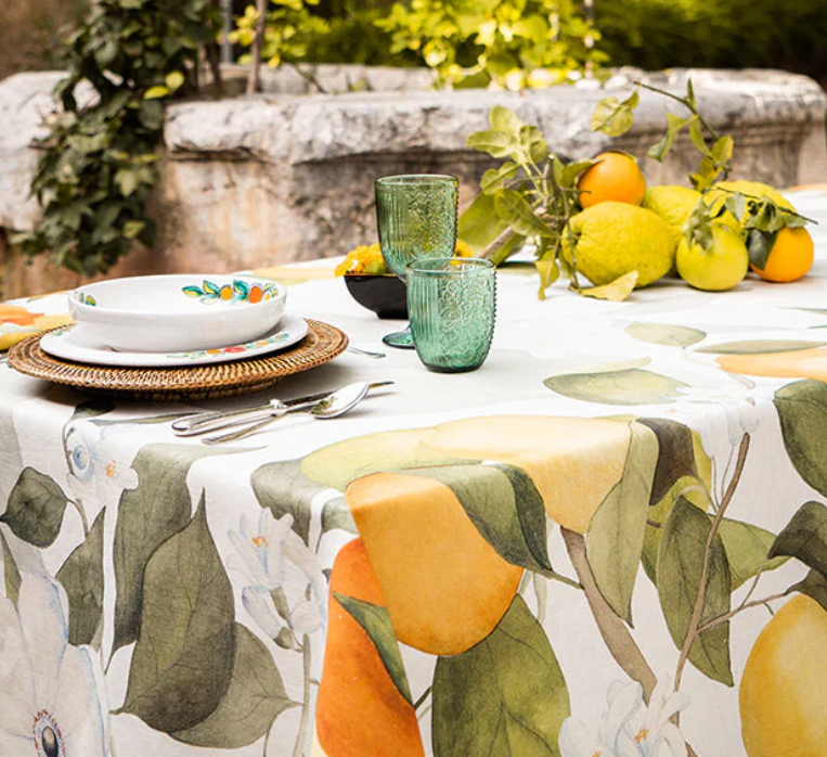 Auf einem Tisch im Freien steht die Napking Premium Leinen Tischdecke 'Amalfi' 110 x 110 cm von Napking, gepaart mit grünen Gläsern, einer weißen Blumenschale, Silberbesteck, Zitrusfrüchten und Grünzeug, vor einer Steinwand und Pflanzen.