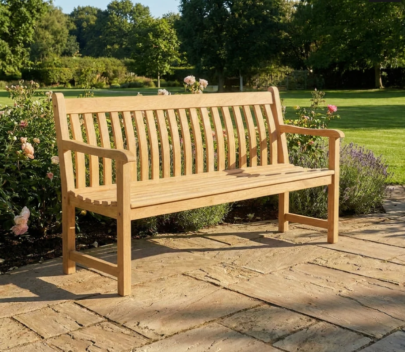Eine Alexander Rose 'Roble Wood Broadfield Bench' Gartenbank 122 cm steht auf einer Steinterrasse in einem sonnenbeschienenen Garten, umgeben von blühenden Blumen, üppigem Gras und Bäumen.