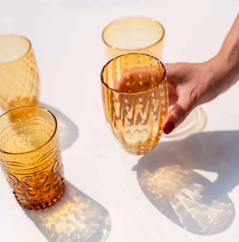 Eine Hand mit rotem Nagellack nimmt ein „Zafferano | Perle Glas Amber 32 cl“ aus einer Gruppe dekorativer bernsteinfarbener Gläser von Zafferano auf einer weißen Fläche in die Hand und wirft dabei gemusterte Schatten, die die exquisite italienische Glasmacherkunst und den einzigartigen, mundgeblasenen Charakter jedes einzelnen Stücks unterstreichen.
