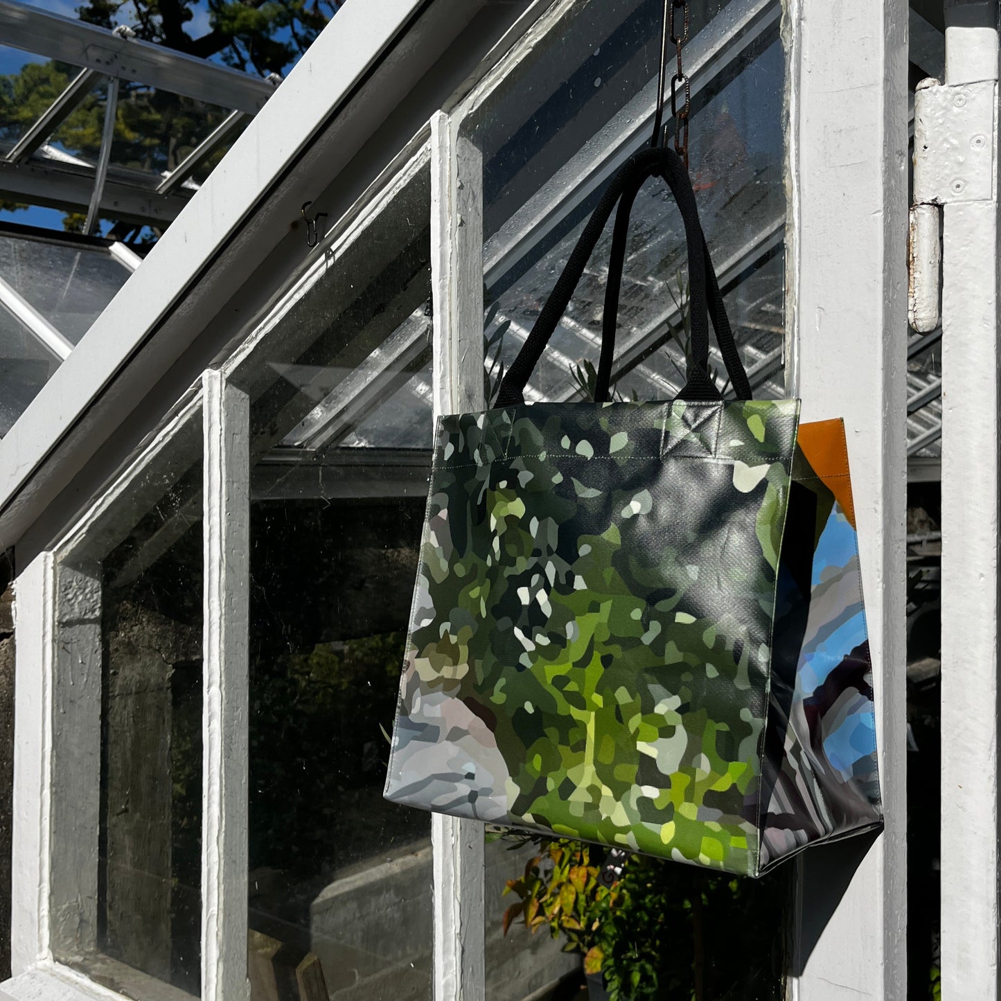 Die Upcycling-Tasche 'Gewächshaus' aus Werbeplane von Königliche Gartenakademie mit einem abstrakten, von der Natur inspirierten Design hängt im Sonnenlicht an einer weiß gerahmten Glastür, aus der Gewächshaus und Grünpflanzen zu sehen sind.