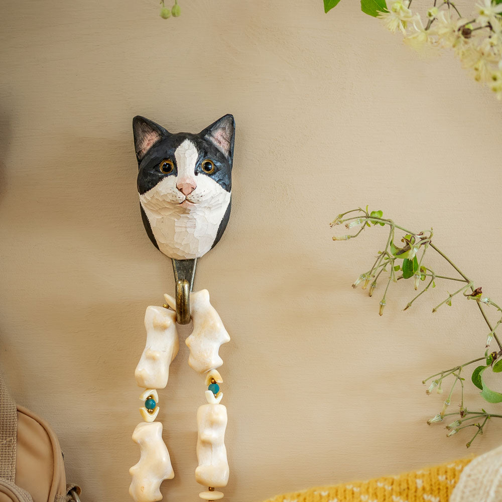 Der handgeschnitzte Wildlife Garden Holz-Wandhaken 'Tuxedo Katze' von Wildlife Garden zeigt einen schwarz-weißen Katzenkopf mit türkisfarbenen Perlen und Knochenanhängern, dekorativ an einer beigen Wand mit grünen Pflanzen platziert.