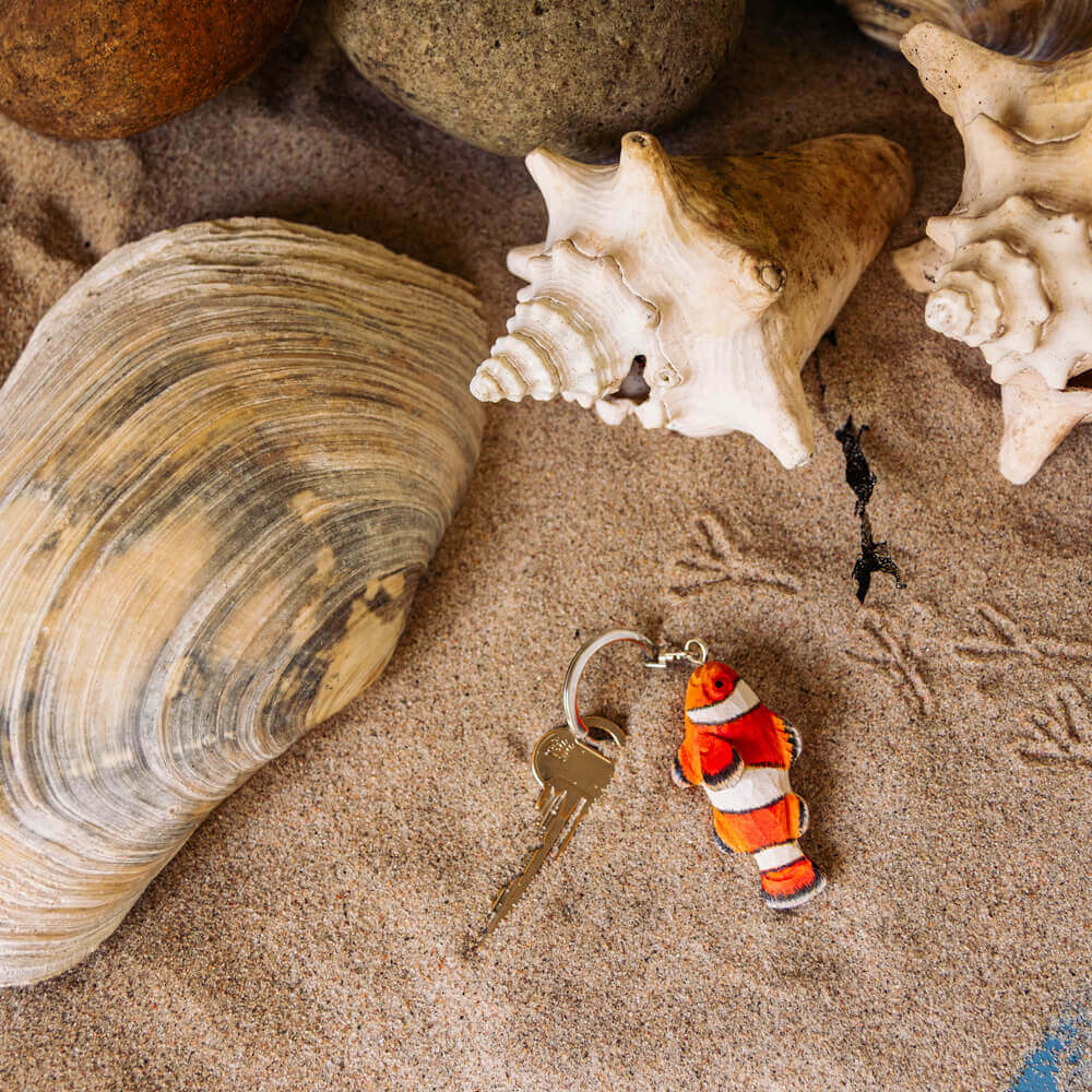 Ein Wildlife Garden Schlüsselanhänger aus Holz, Clownfisch-Design, liegt auf sandigem Boden mit Muscheln und Steinen - Vogelspuren im Sand machen ihn zum charmanten Geschenk für Meeresliebhaber.