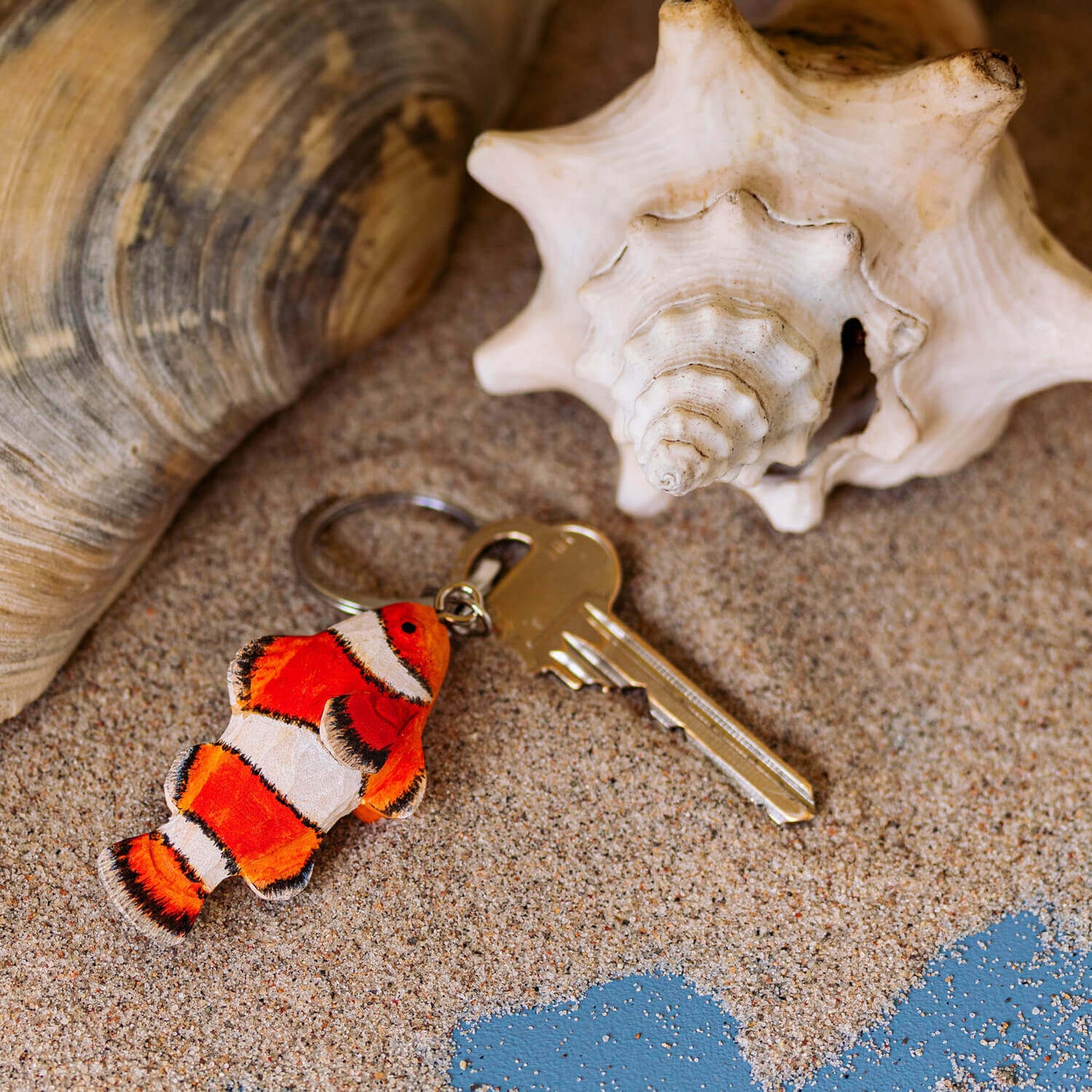 Ein handgefertigter Schlüsselanhänger aus Holz | Clownfisch von Wildlife Garden liegt neben Muscheln im Sand - ein charmantes Geschenk für Meereswelt-Fans.