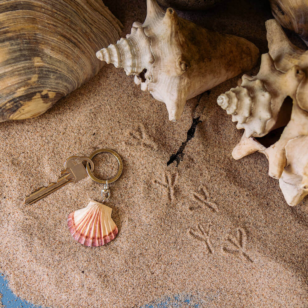 A Wildlife Garden Handgefertigter Schlüsselanhänger aus Holz Muschel liegt auf sandigem Boden neben großen Muscheln und Vogelspuren - ein charmantes maritimes Accessoire von Wildlife Garden.