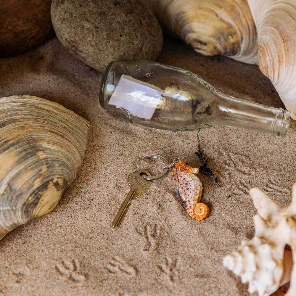 A Wildlife Garden Handgefertigter Schlüsselanhänger aus Holz in Seepferd-Form liegt auf Sand neben einer Flasche, Muscheln und Spuren-ein maritimes Accessoire der Marke Wildlife Garden, ideal für Strandfans.