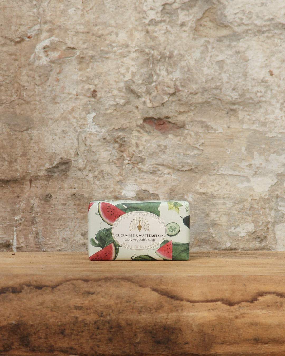 Eine Tafel The English Soap Company ESC | 'Wassermelone & Gurke' erfrischend fruchtige pflanzliche Handseife, in thematischer Verpackung, ruht auf rustikalem Holz vor einer strukturierten Steinwand.