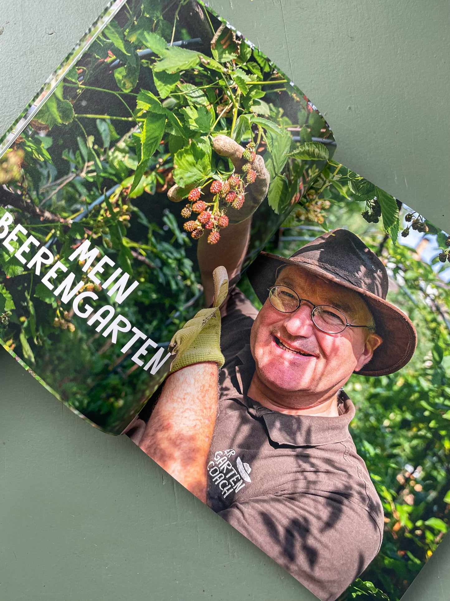Ein lächelnder Mann mit Hut, Brille, Handschuhen und einem braunen Hemd steht in einem Beerengarten und pflückt reife Beeren. Auf der linken Seite steht "MEIN BEERENGARTEN". Produkt: Willkommen beim Gartencoach | Signierte Ausgabe von Edition Lempertz.