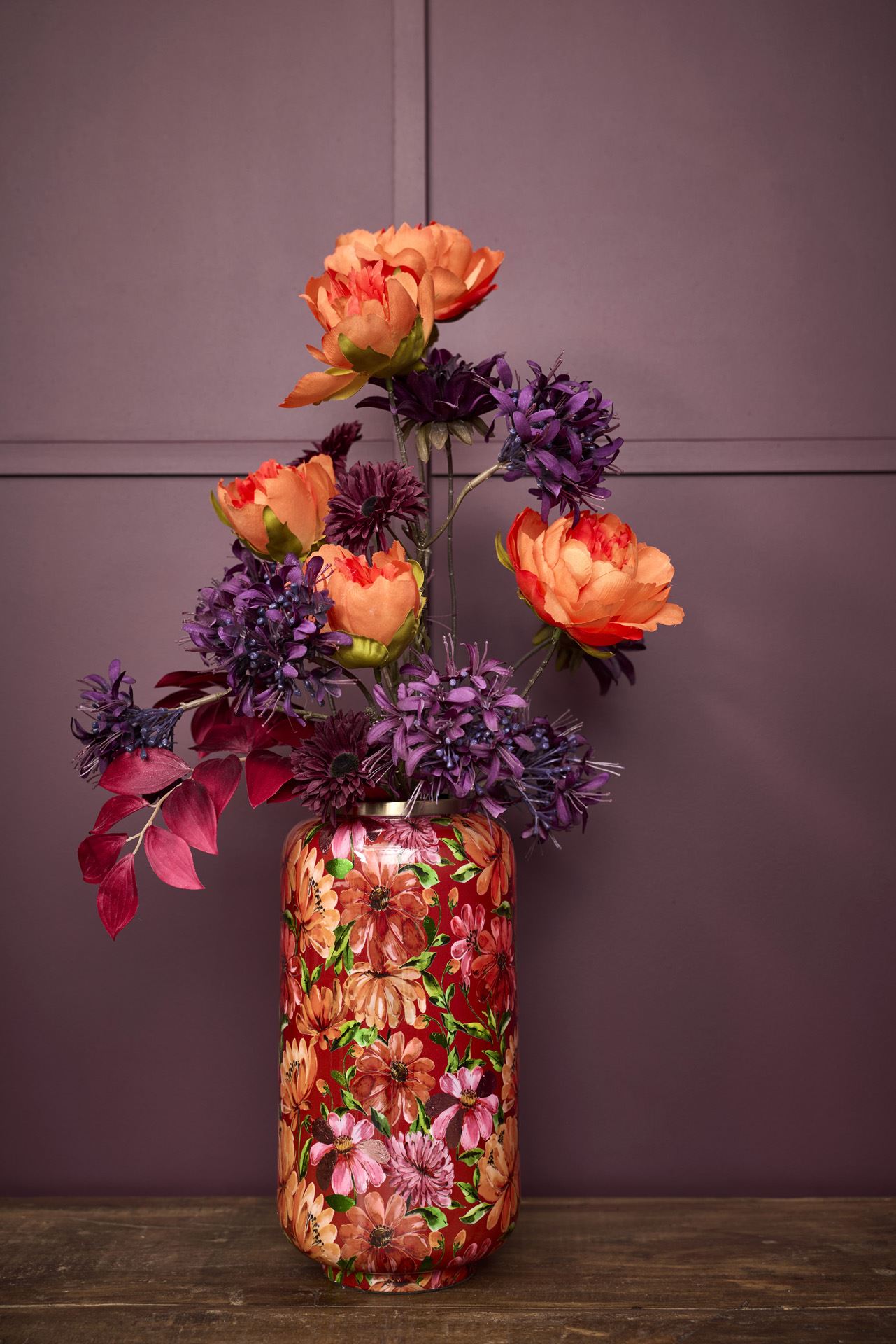 Ein farbenfrohes Blumenarrangement mit orangefarbenen und violetten Blüten ist in der Vase „Bloom“ aus Metall in Rosarot (30 x 15 cm) von 2 Have zu sehen, die sich durch ein dekoratives Blumenmuster auszeichnet. Die Vase steht auf einer Holzunterlage vor einer violetten Wand.