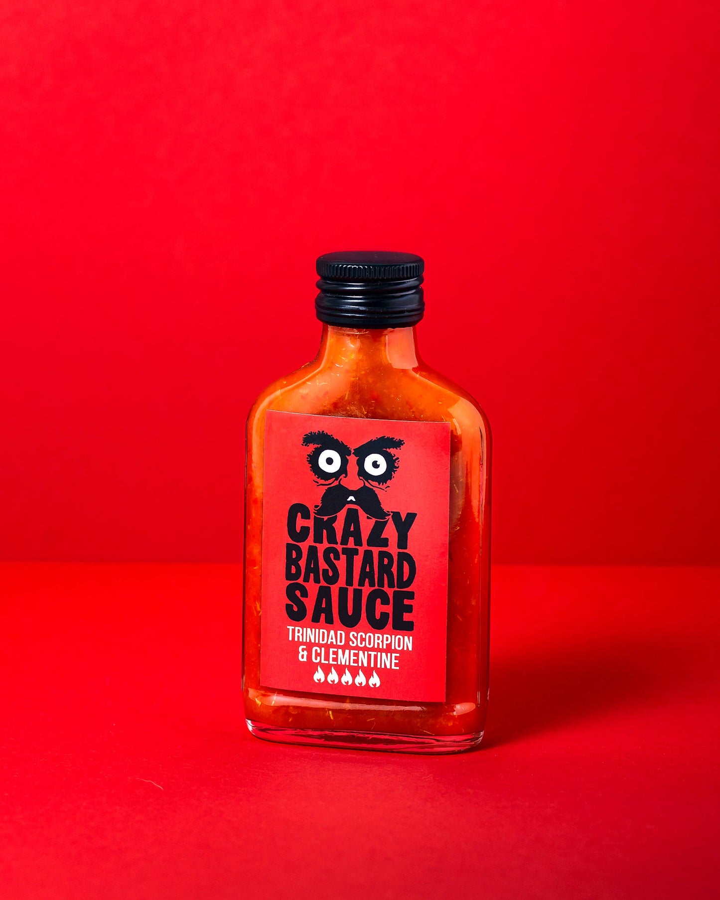 Eine quadratische Glasflasche mit Crazy Bastard Sauce Trinidad Scorpion & Clementine 100 ml scharfe Soße steht auf einer roten Fläche mit rotem Hintergrund. Das Etikett zeigt stilisierte Augen, Text und Schärfesymbole für Chilischoten am unteren Rand.