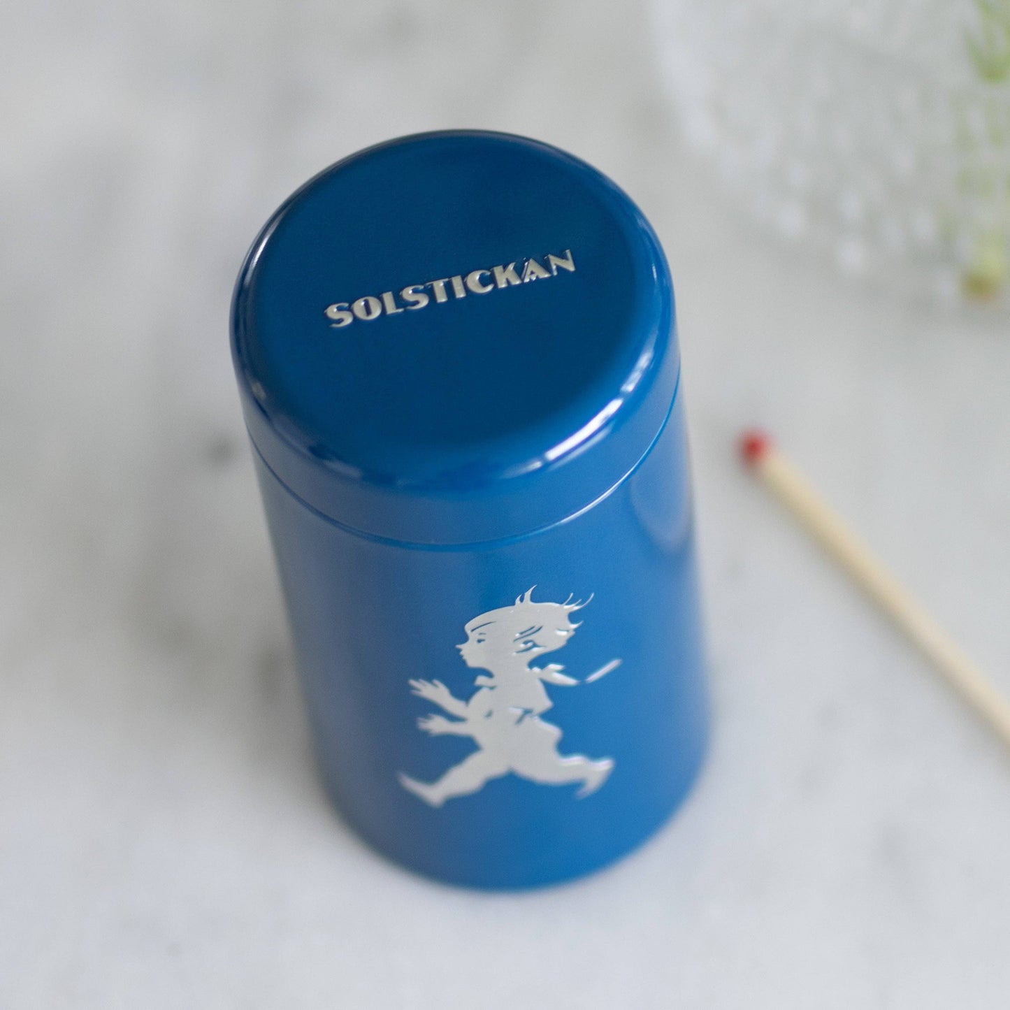 Eine Solstickan Design | Streichholzdose Blau, eine blaue zylindrische Streichholzschachtel von Solstickan Design mit der weißen Silhouette eines laufenden Kindes und SOLSTICKAN auf dem Deckel, wird auf einer hellen Fläche neben einem Holzstreichholz gezeigt.