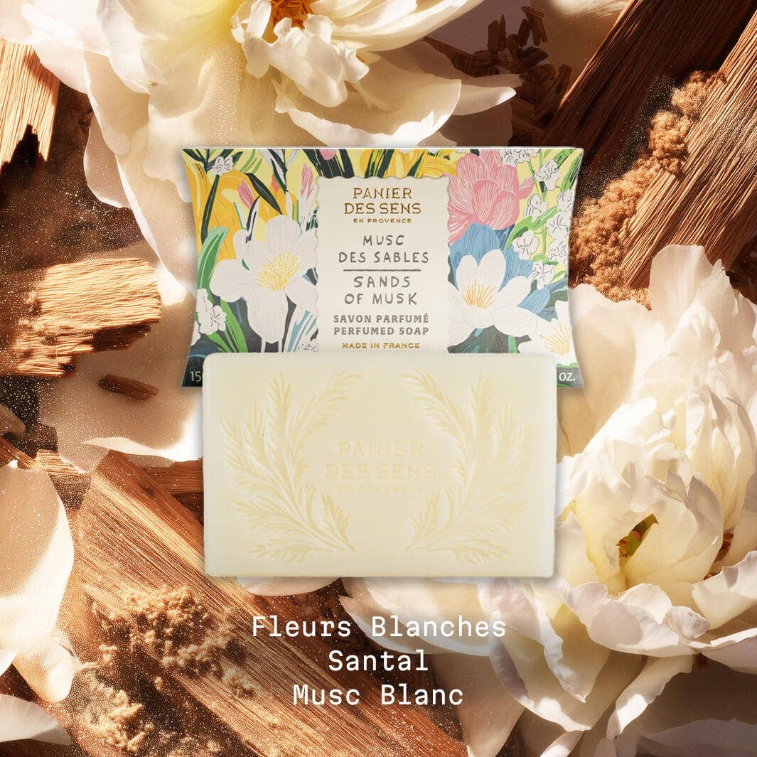 Der Panier Des Sens | Duftseife Musc des Sables 150 g ruht auf cremigen Blütenblättern mit seiner dekorativen Schachtel dahinter. Fleurs Blanches, Santal, Musc Blanc. Perfekt als festliche Badeseife für Ihr Ritual.
