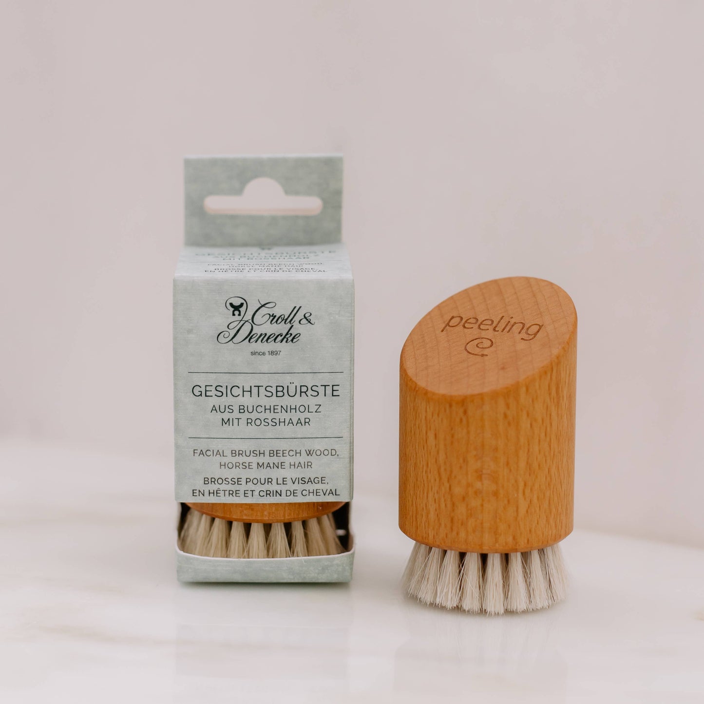 Eine Croll & Denecke Gesichtsbürste aus Holz für Peeling & Massage mit hellen Rosshaarborsten steht neben ihrer Schachtel, die in Deutsch und Englisch beschriftet ist. Auf dem Griff aus Buchenholz ist "Peeling" aufgedruckt.