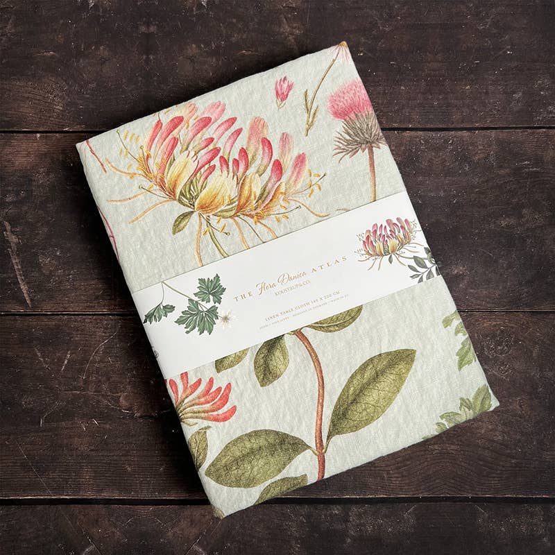 Eine gefaltete Tischdecke von Koustrupco | „Flora Danica Atlas“ (350 × 145 cm), verziert mit botanischen Illustrationen von Blüten und Blättern und mit einem Papierband mit der Aufschrift „The Flora Danica Atlas“ umwickelt, liegt auf einer dunklen Holzfläche.