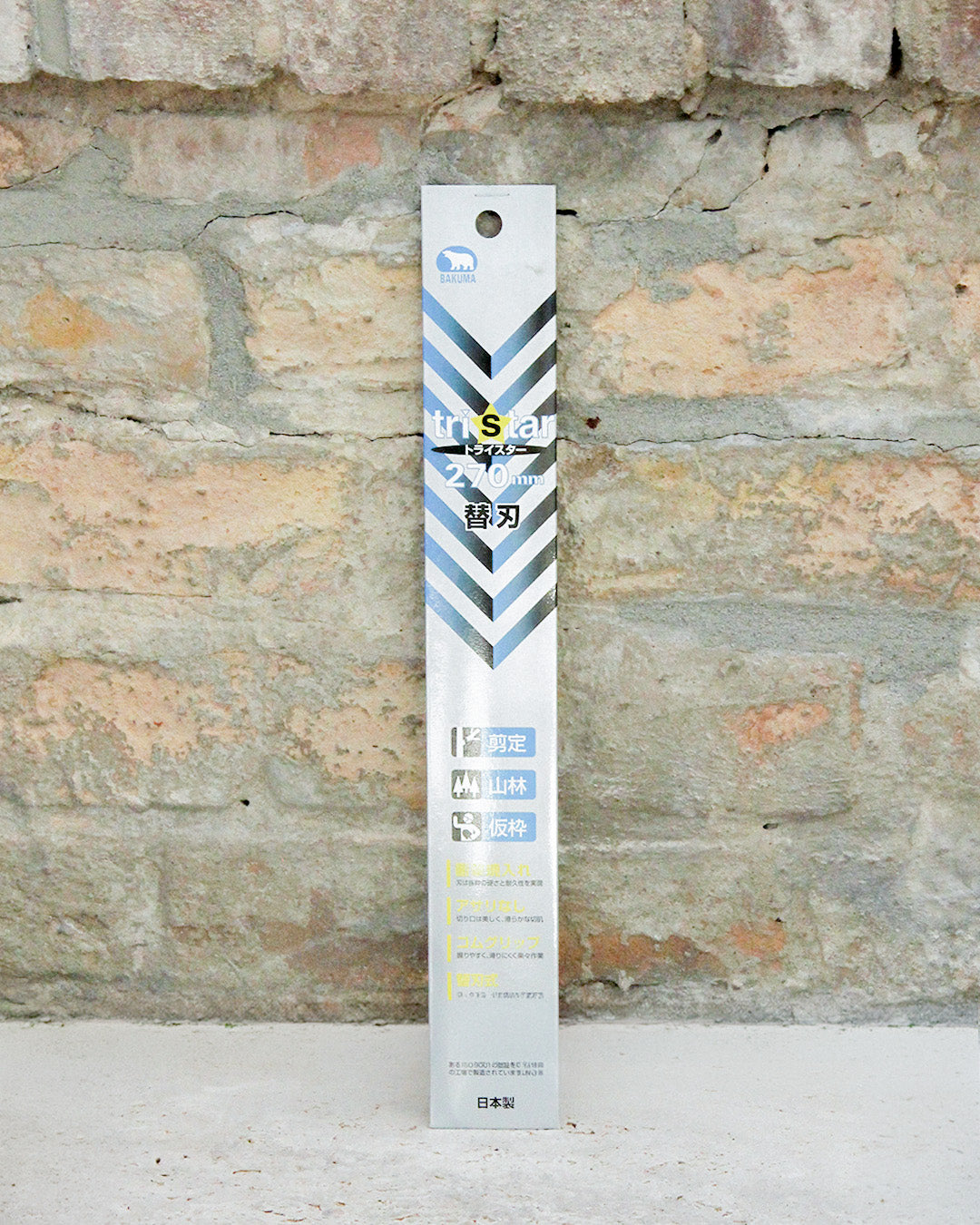 Hohe, rechteckige silberne Schachtel mit blauen Chevron-Streifen, die aufrecht vor einer rauen Backsteinmauer steht. Auf der Verpackung sind japanische und englische Texte zu sehen, darunter der Markenname „Niwaki“ und der Produktname „Ersatzsägeblatt für Niwaki GR 270“.