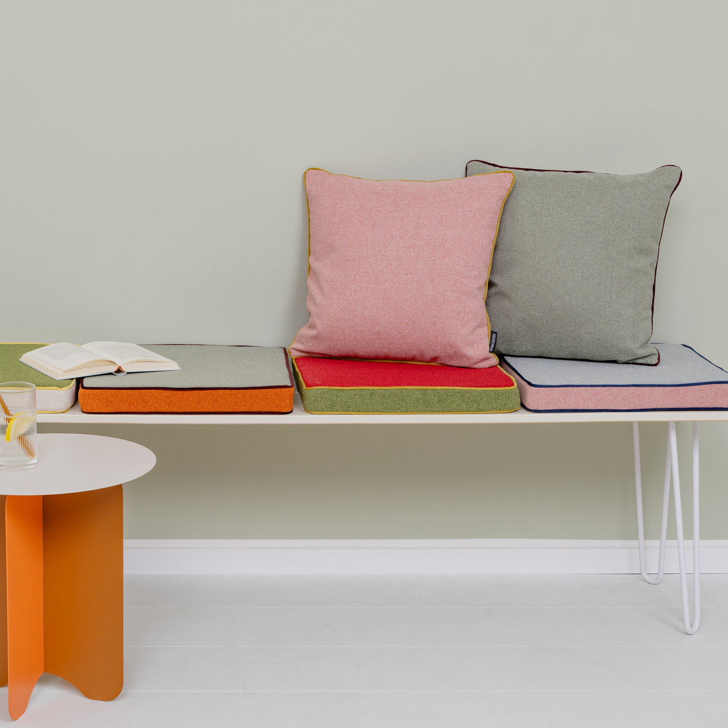 Eine minimalistische Szene mit einer weißen Bank mit dem Remember Kissen 'Cranberry' (45 x 45 cm) als gemütliches Wohnaccessoire. Ein aufgeschlagenes Buch und ein Glas Wasser mit Zitrone stehen auf einem kleinen weiß-orangefarbenen Beistelltisch daneben.