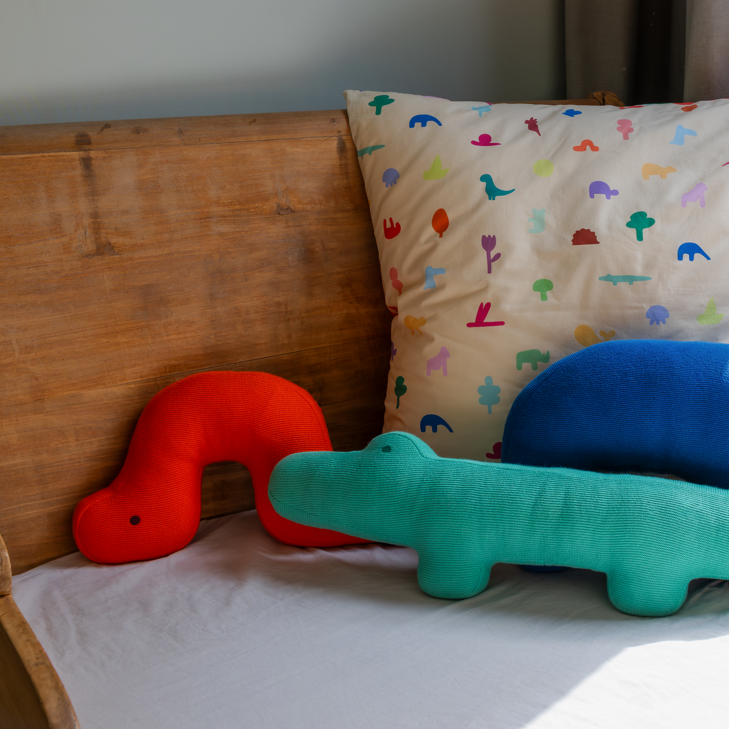 Ein Holzbett mit weißem Laken in einem gemütlichen Kinderzimmer mit zwei Plüschsauriern - einem roten und einem blaugrünen -, einem Remember Kuschelkissen "Ameisenbär Edgar" und Sonnenlicht, das auf das Bett fällt.