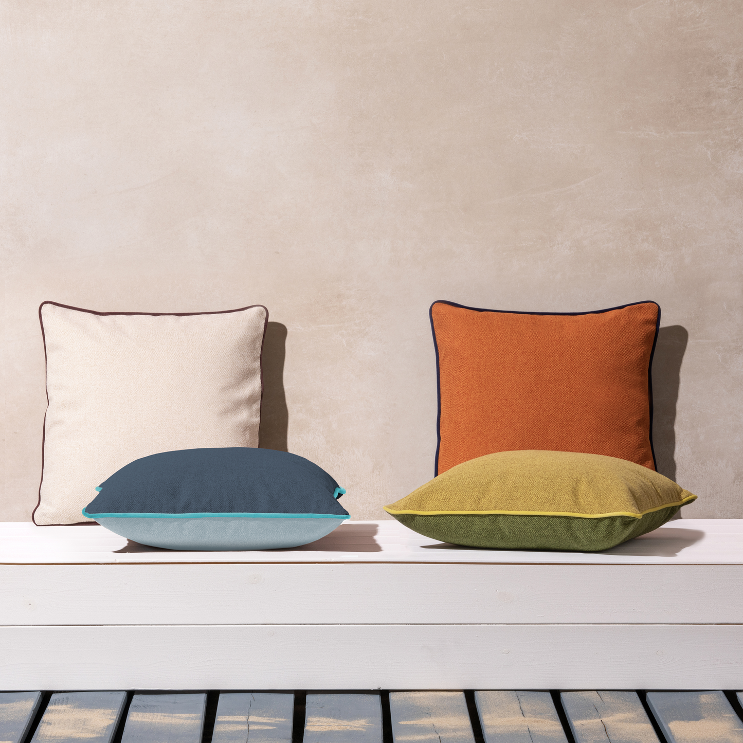 Das Remember Kissen 'Peanut' 45 x 45 cm in Creme, Orange, Blau und Grün mit eleganter Samtpaspelierung verleiht jedem Raum einen stilvollen Touch, besonders auf einer weißen Bank vor einer beigen Wand über einem gestreiften Holzboden.
