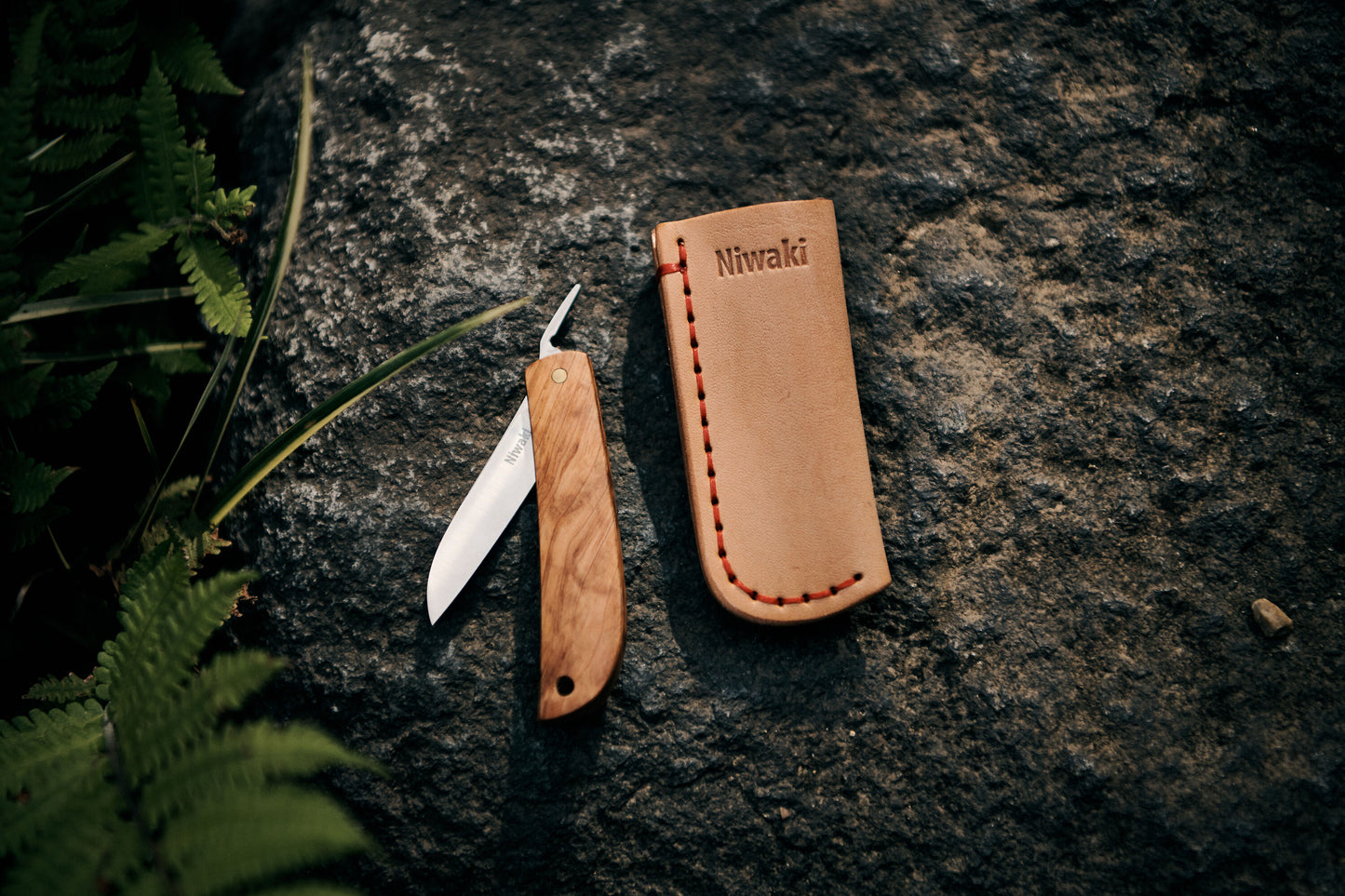 Ein Niwaki | 'Kotoh X Niwaki Knife' Taschenmesser mit Schutzhülle aus Leder liegt mit geöffnetem Holzgriff auf einem Felsen neben der tanfarbenen, Niwaki-gebrandeten Lederschutzhülle, umgeben von grünen Farnen - ideal für Gartenarbeit.